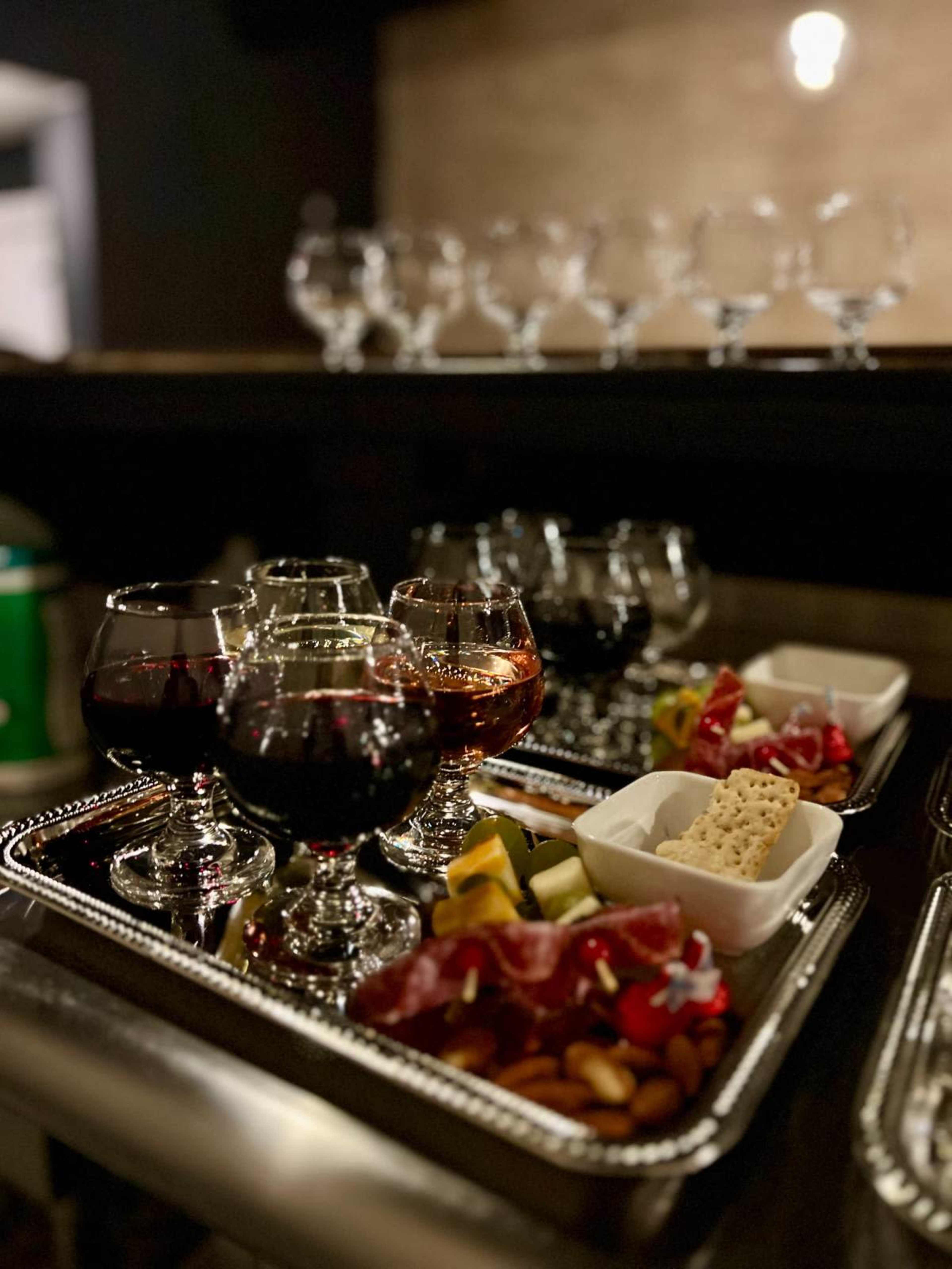 A tray displays several glasses of red and amber liquid alongside assorted meats, cheeses, nuts, and fruits.