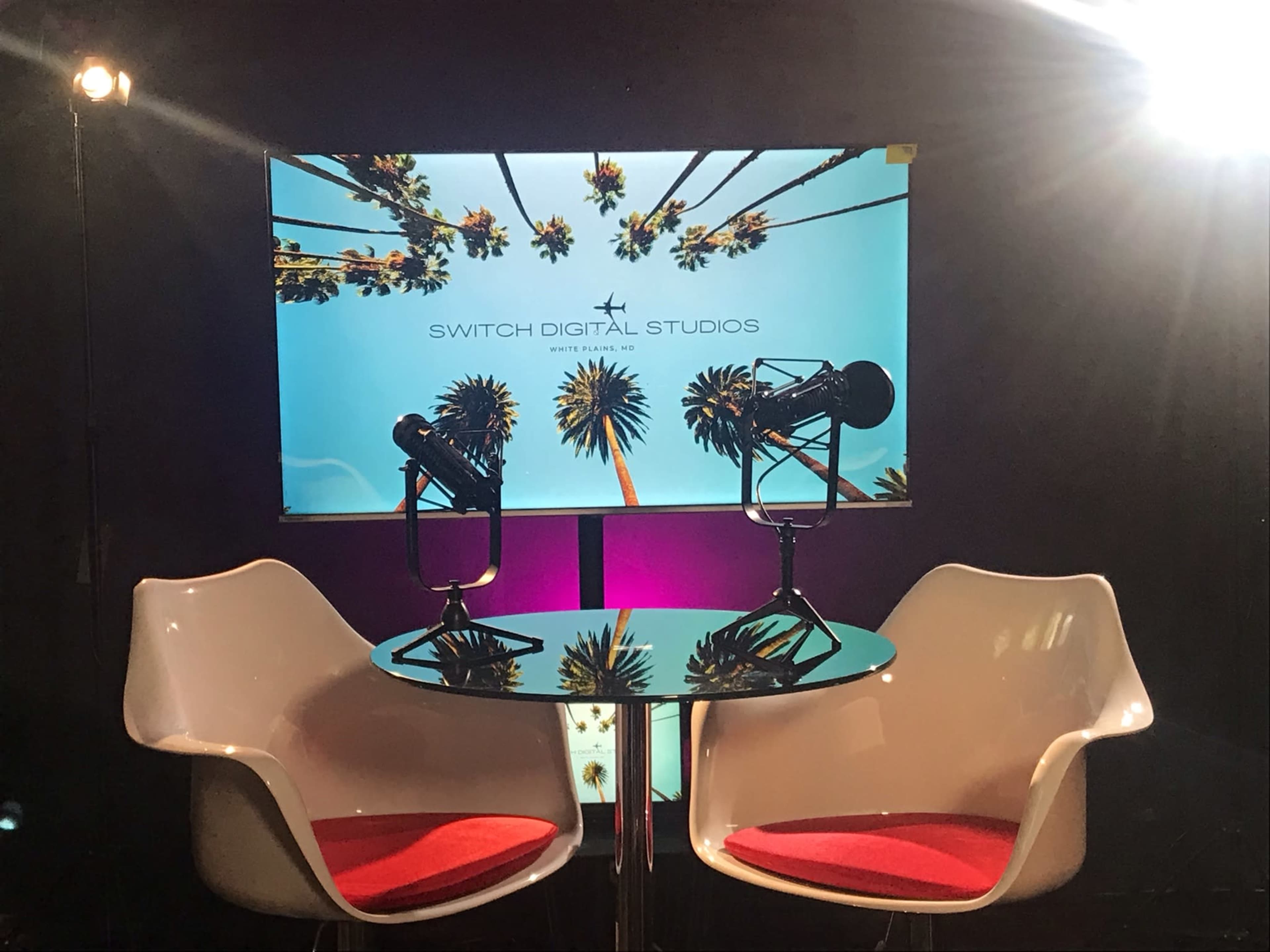 A modern studio setup featuring two white chairs, a circular table, and a backdrop with palm trees and the text "SWITCH DIGITAL STUDIOS."