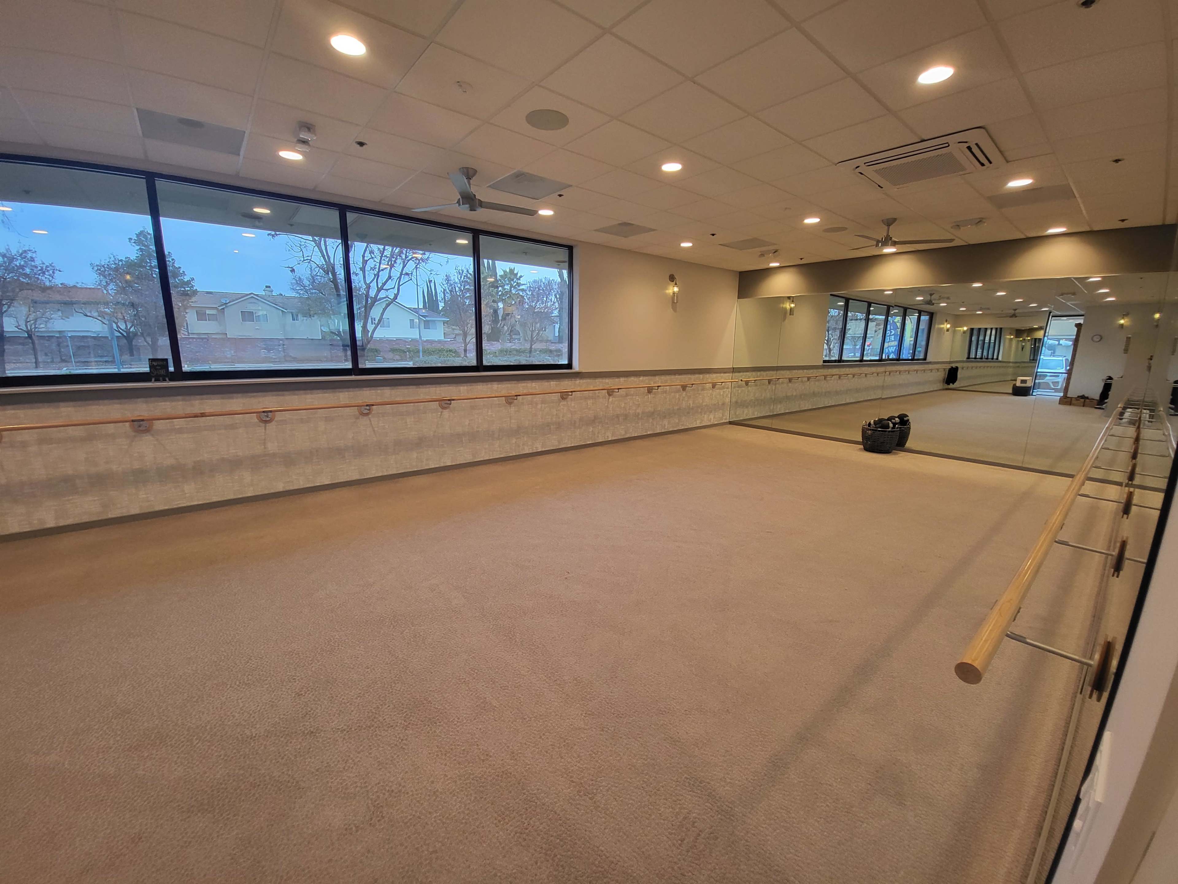 An empty studio space with large windows, a mirrored wall, and a ballet barre along one side.