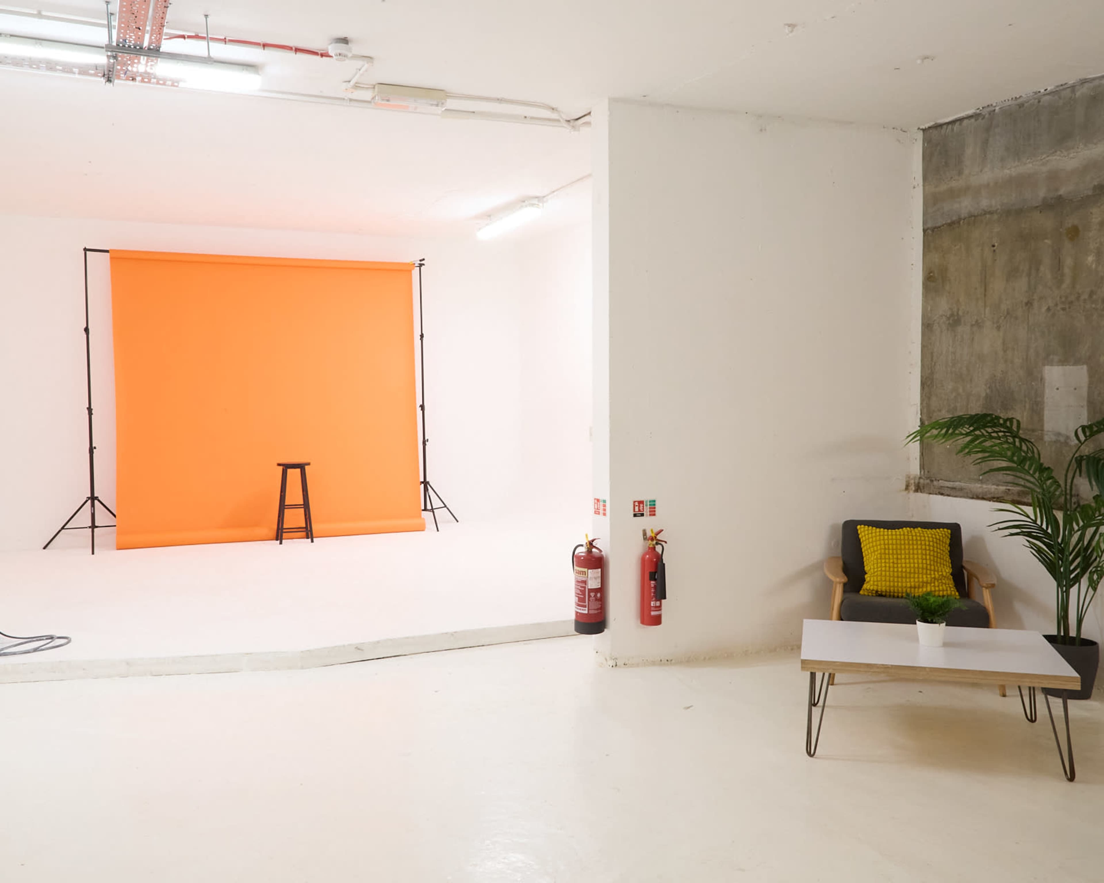 The image shows a bright, empty photography studio with an orange backdrop, a stool, and a seating area featuring a plant and a coffee table.