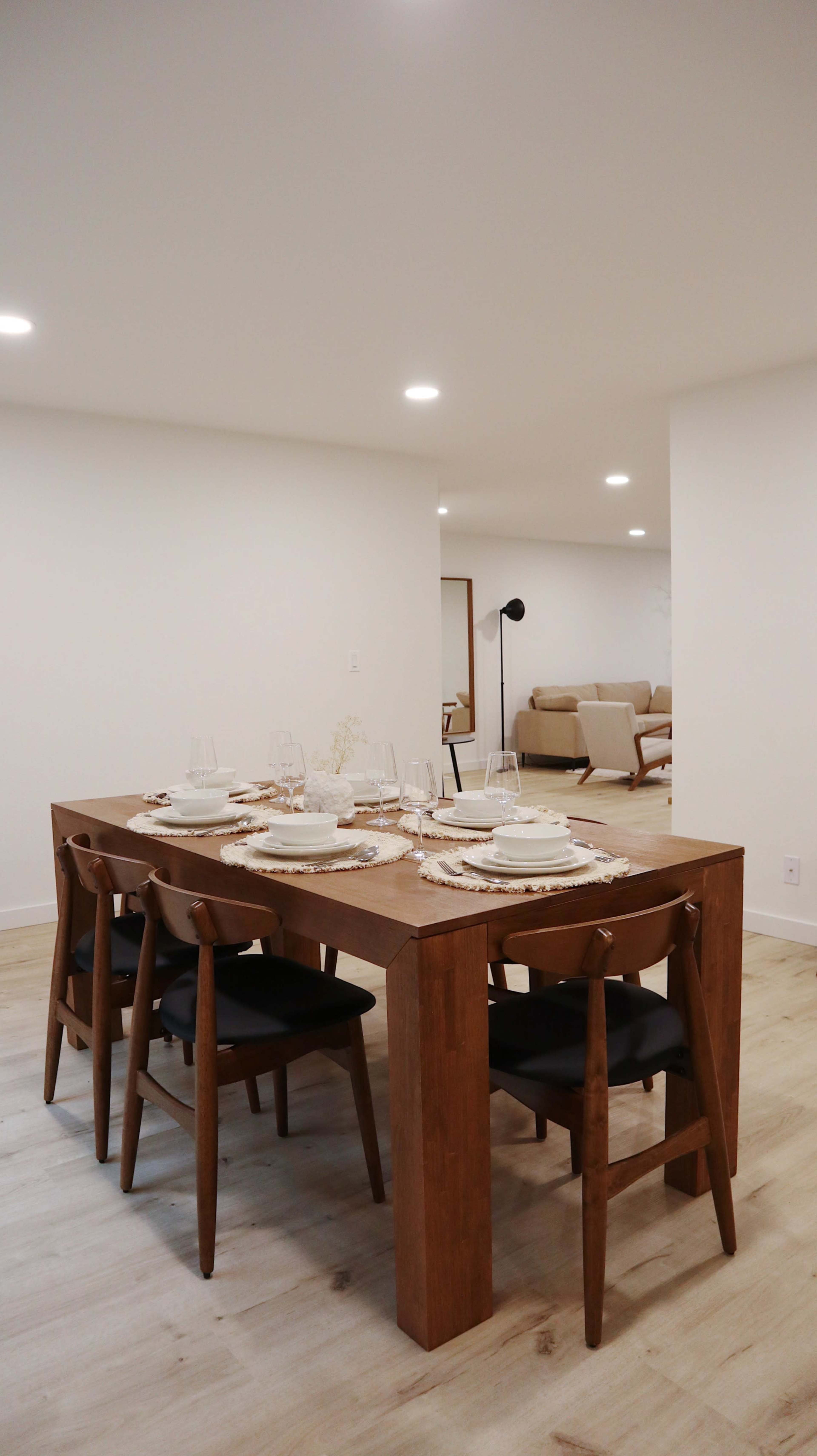 A wooden dining table is set with dishes and glasses, surrounded by chairs, while a living area is visible in the background.