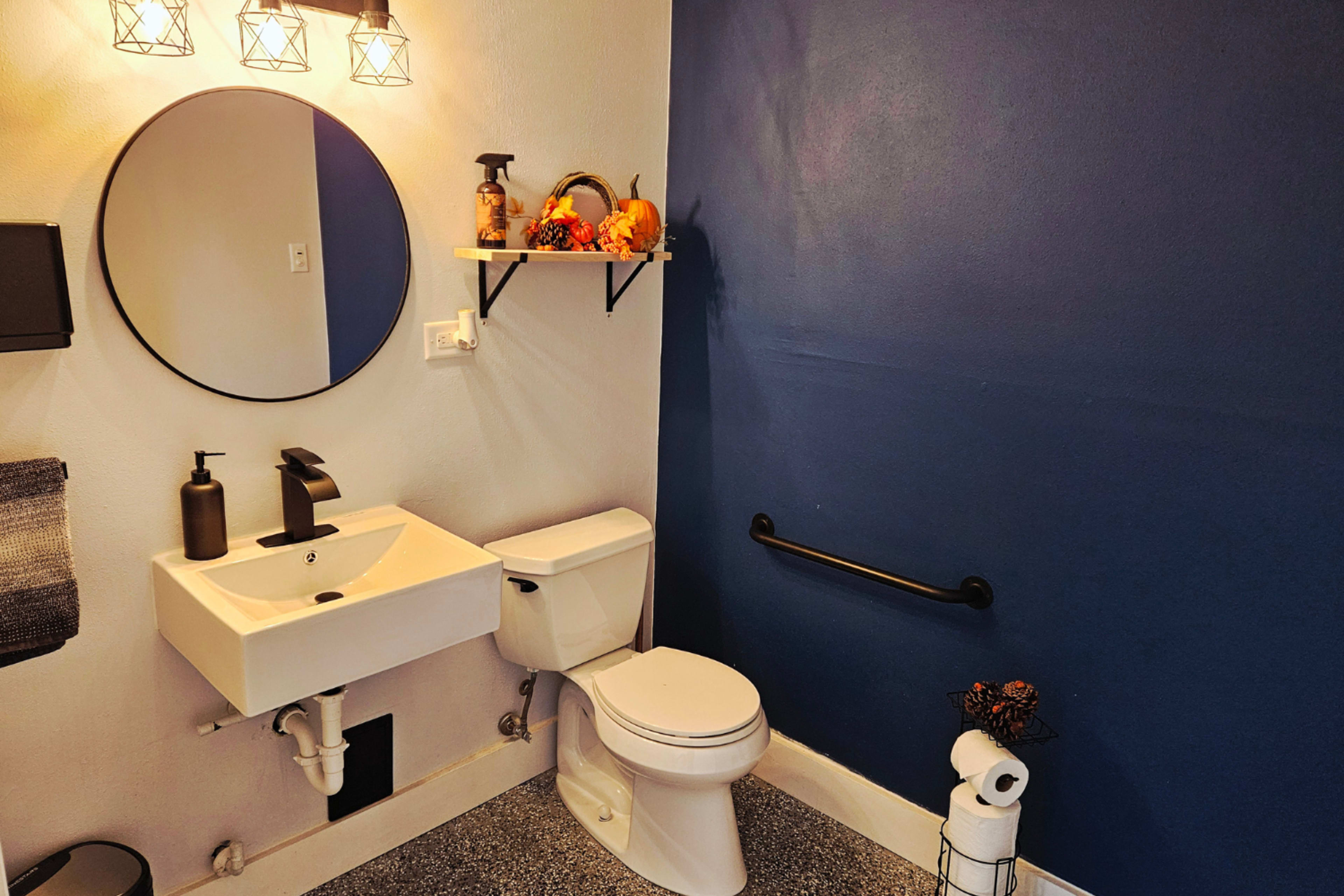 The image shows a small bathroom featuring a round mirror above a modern sink, a toilet, and a wall-mounted shelf with decorative items, against a dark blue wall.