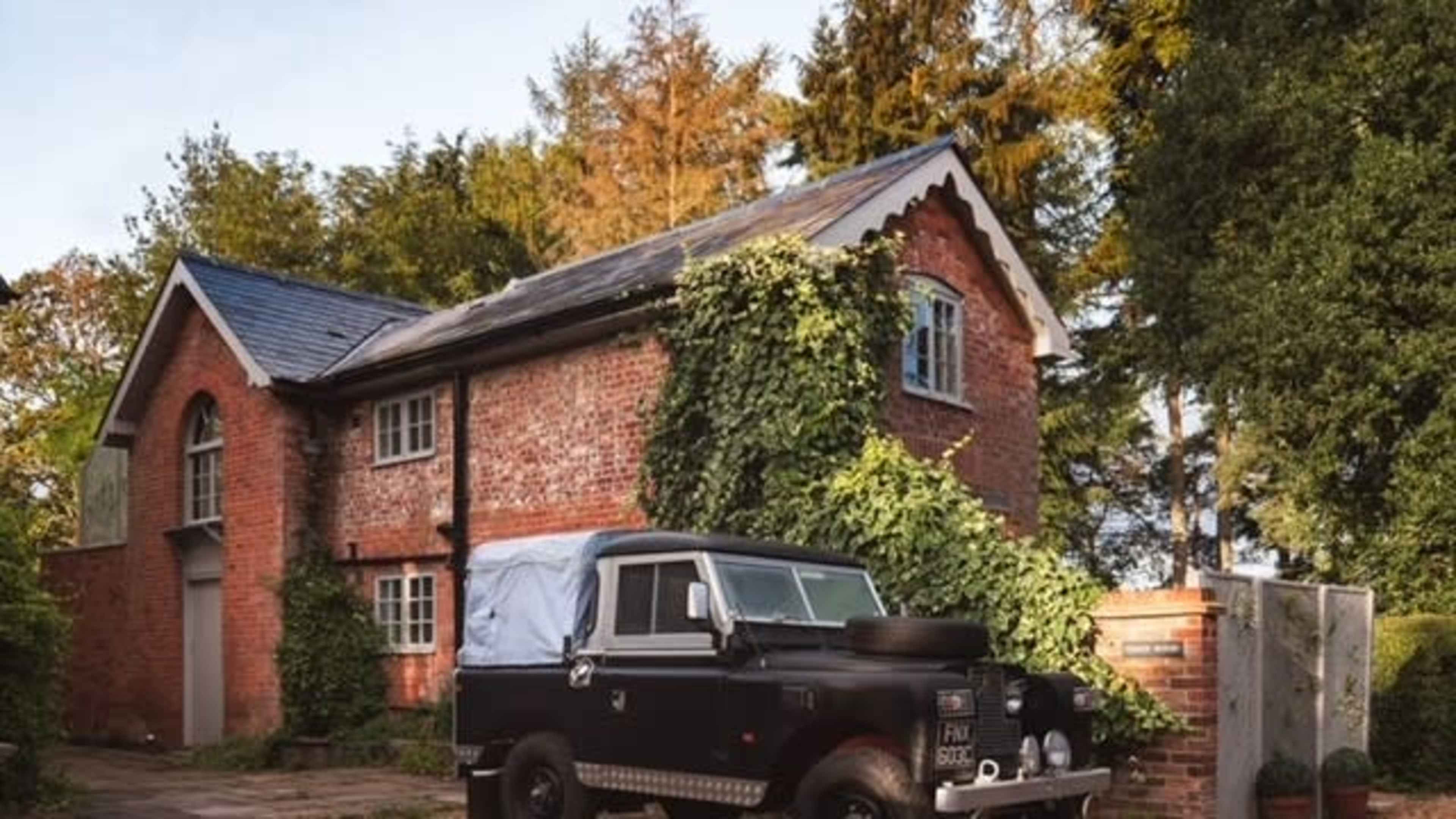 An old black truck is parked in front of a brick building covered with greenery in a wooded area.
