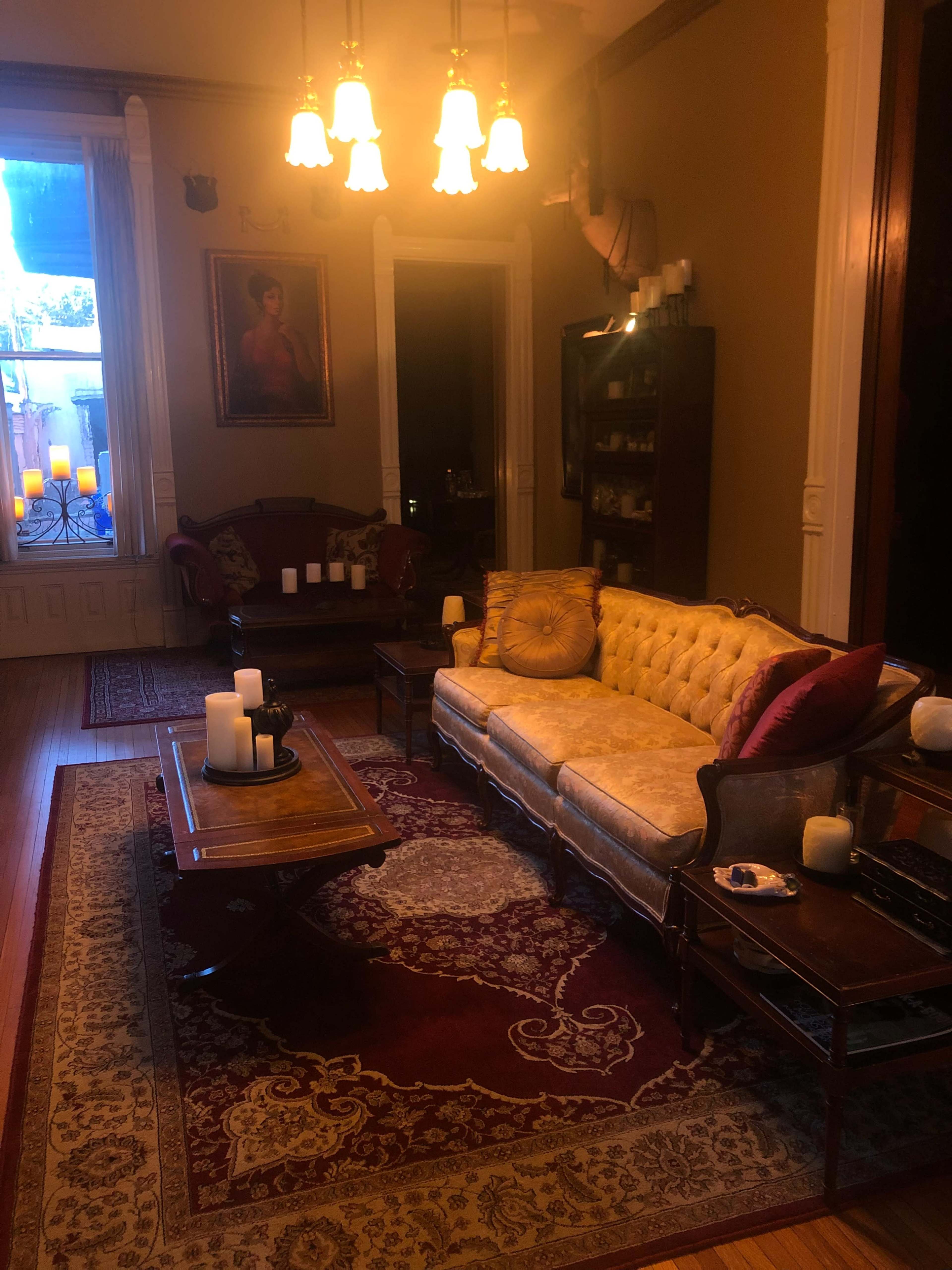 A dimly lit living room with a vintage couch, a wooden coffee table, and multiple candles arranged on furniture, set against a backdrop of ornate decor.