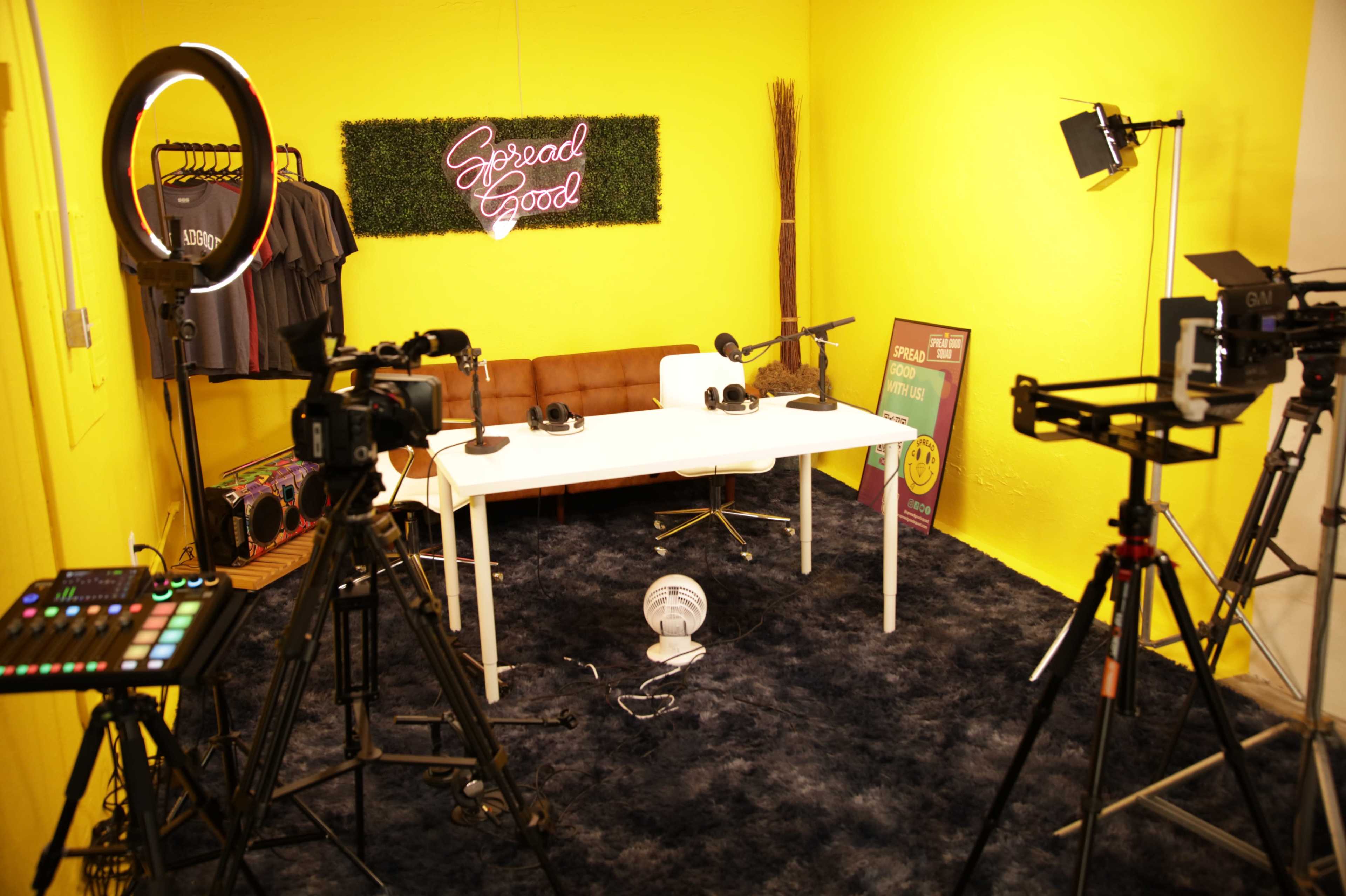 The scene shows a podcast studio with a yellow wall, a white table, microphones, headphones, and various cameras set up around the room.