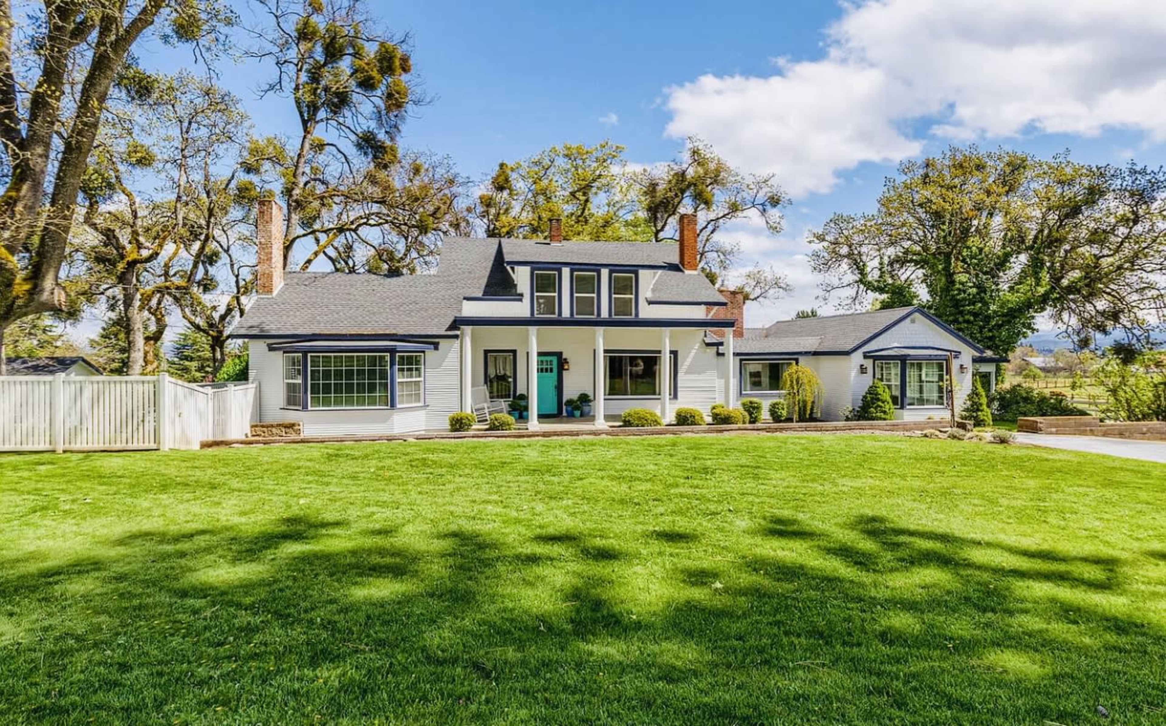 A two-story house with a gable roof and large windows is set on a green lawn surrounded by trees and shrubs.