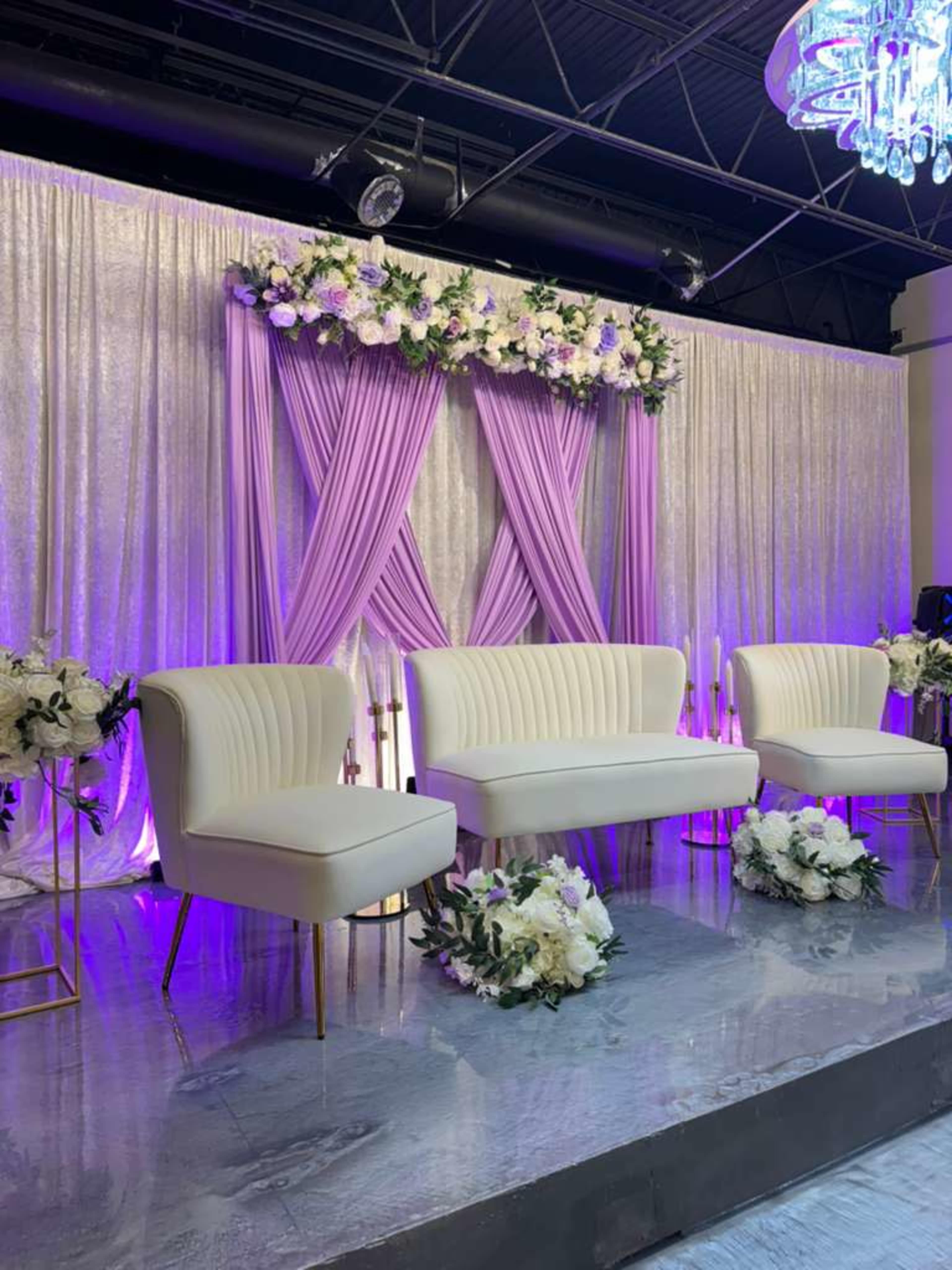 The image shows a decorated stage with a white backdrop, purple drapery, and floral arrangements, featuring a set of white chairs.