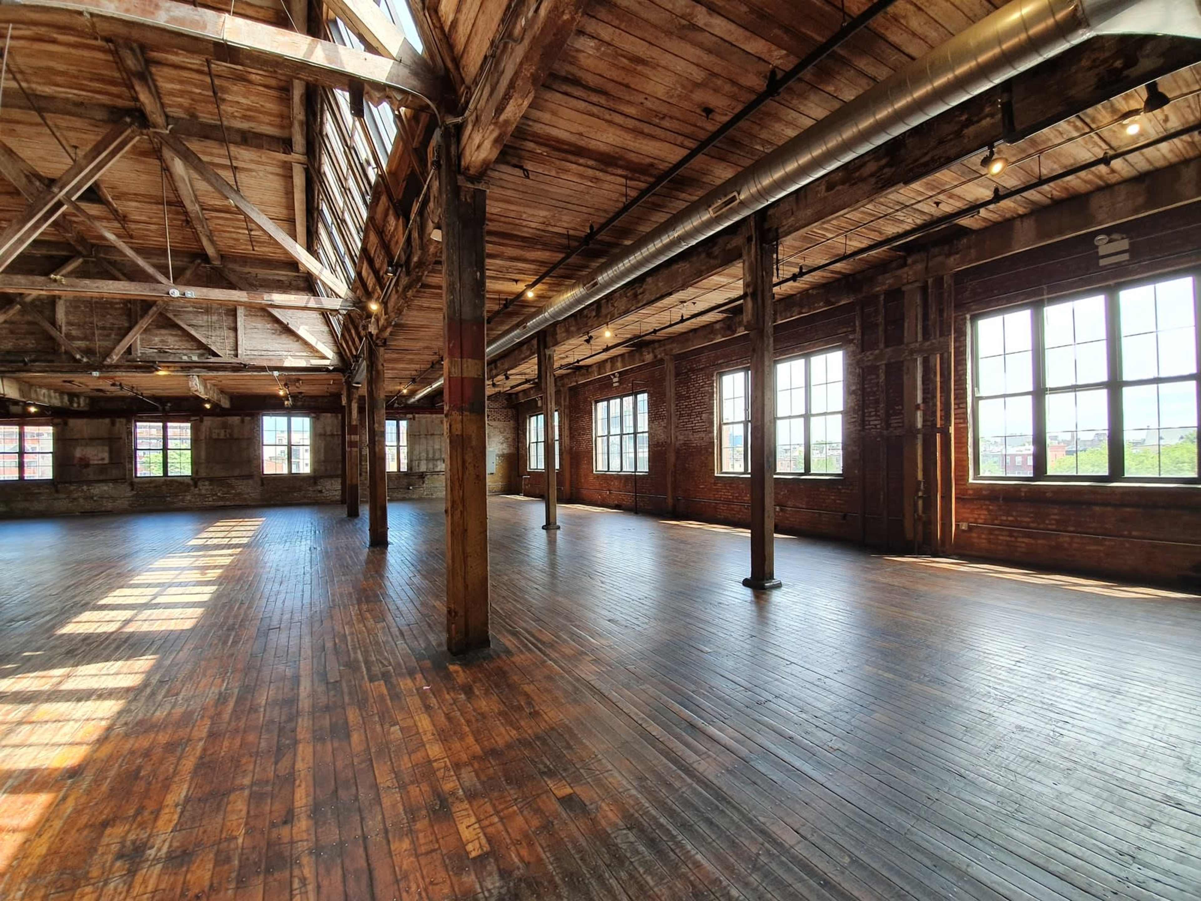 A spacious, empty warehouse interior with wooden beams and large windows casting light on the hardwood floor.