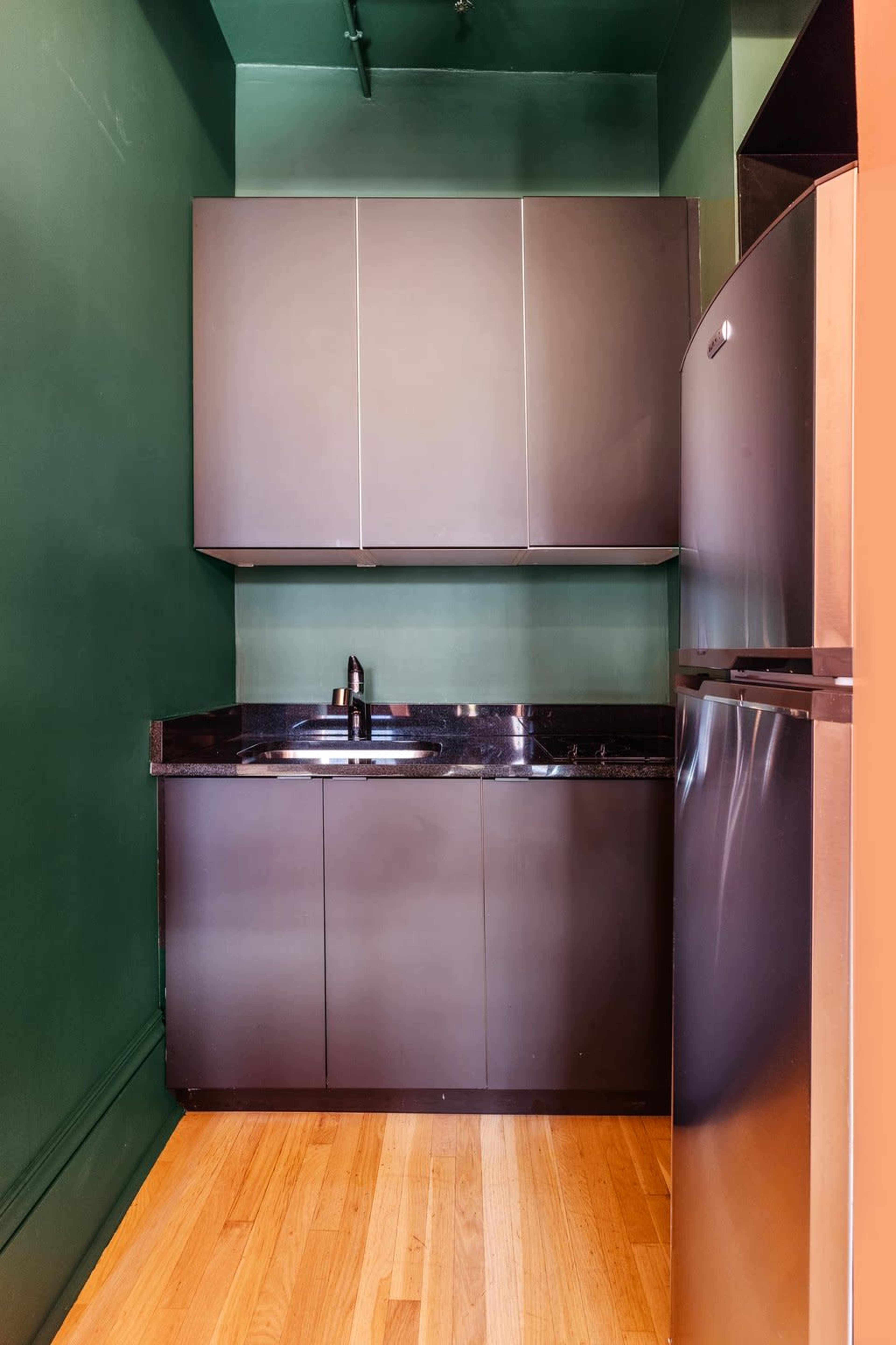 A compact kitchen with green walls, a stainless steel refrigerator, a black countertop, and gray cabinetry.