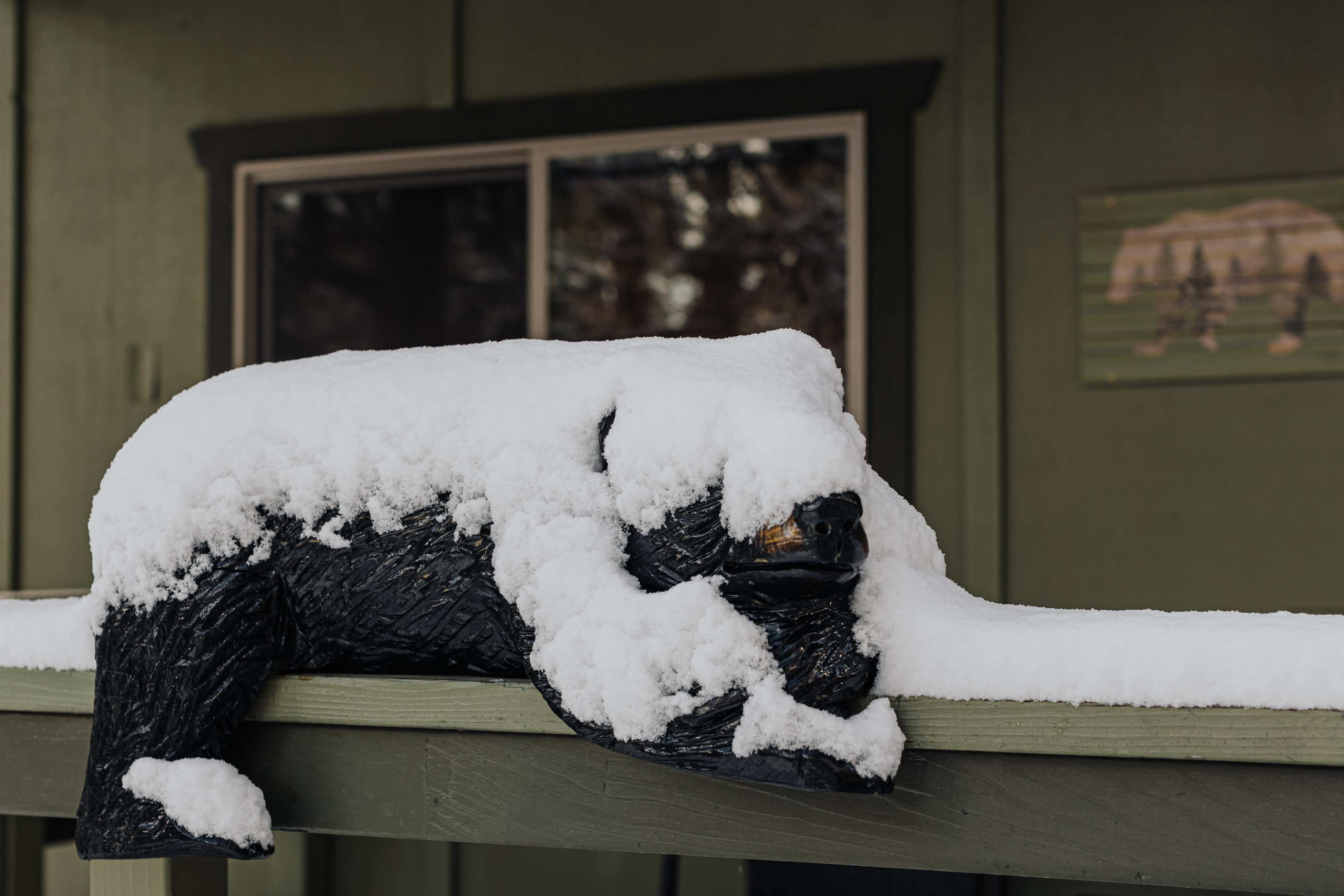 A black bear statue sits on a wooden railing, covered in a thick layer of snow.