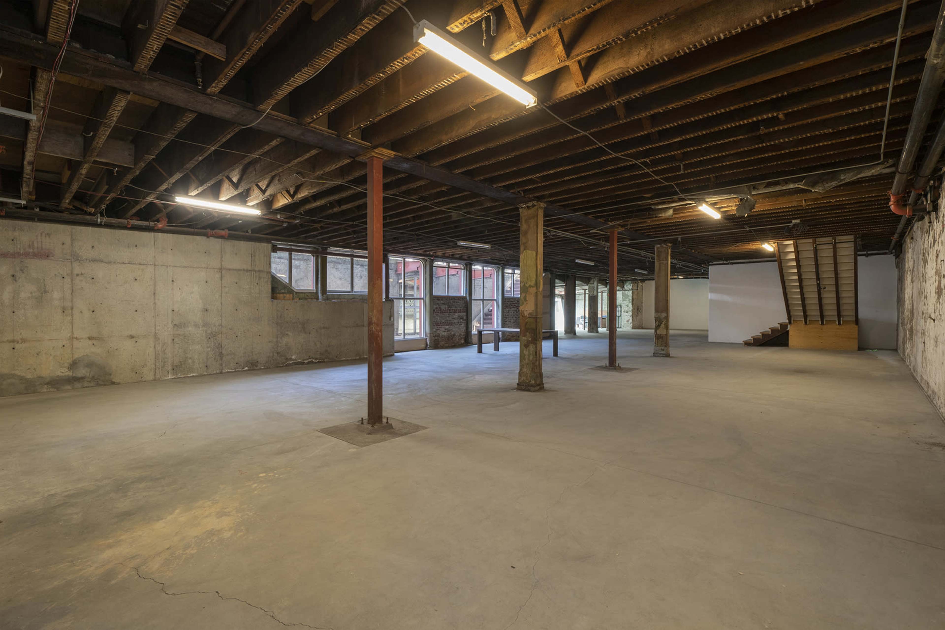A spacious, unfinished basement with exposed beams, concrete walls, and a staircase at one end.