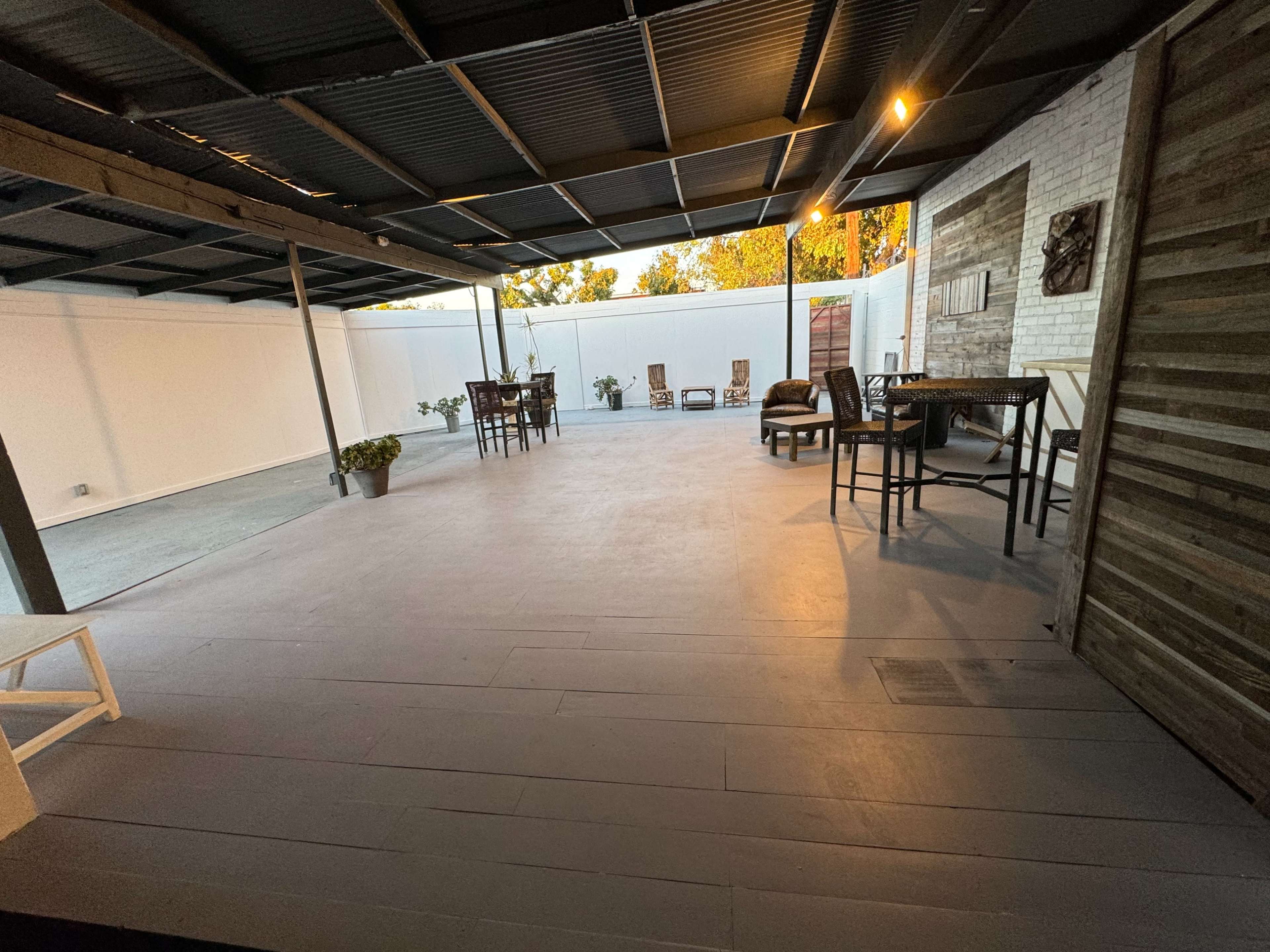 The image shows a spacious outdoor patio area with a shaded roof, featuring several tables and chairs, and surrounded by white walls and plants.