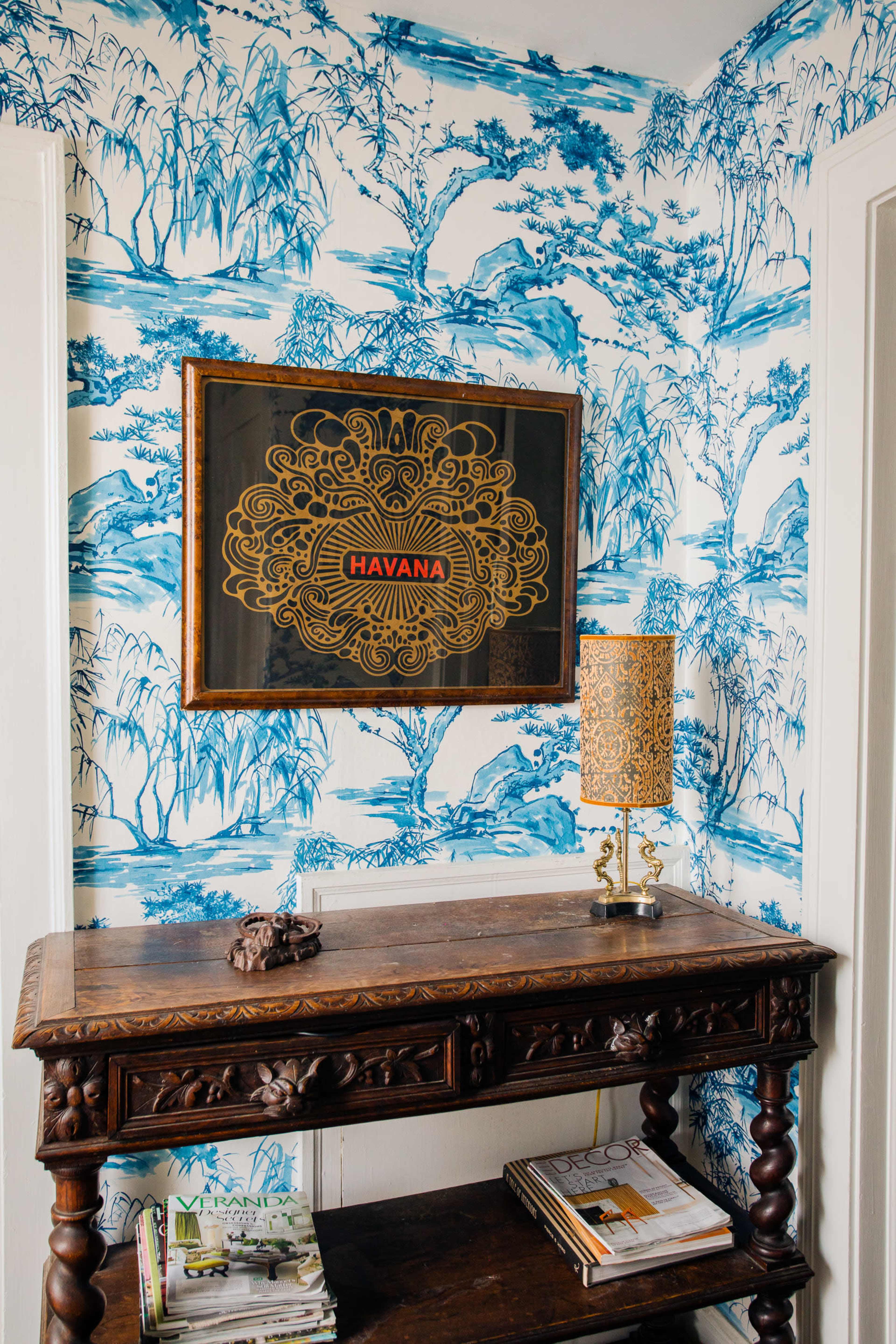 A wooden table with magazines rests against a patterned wallpaper featuring blue landscapes, with a gold-framed artwork labeled "HAVANA" above it and a decorative lamp on the table.