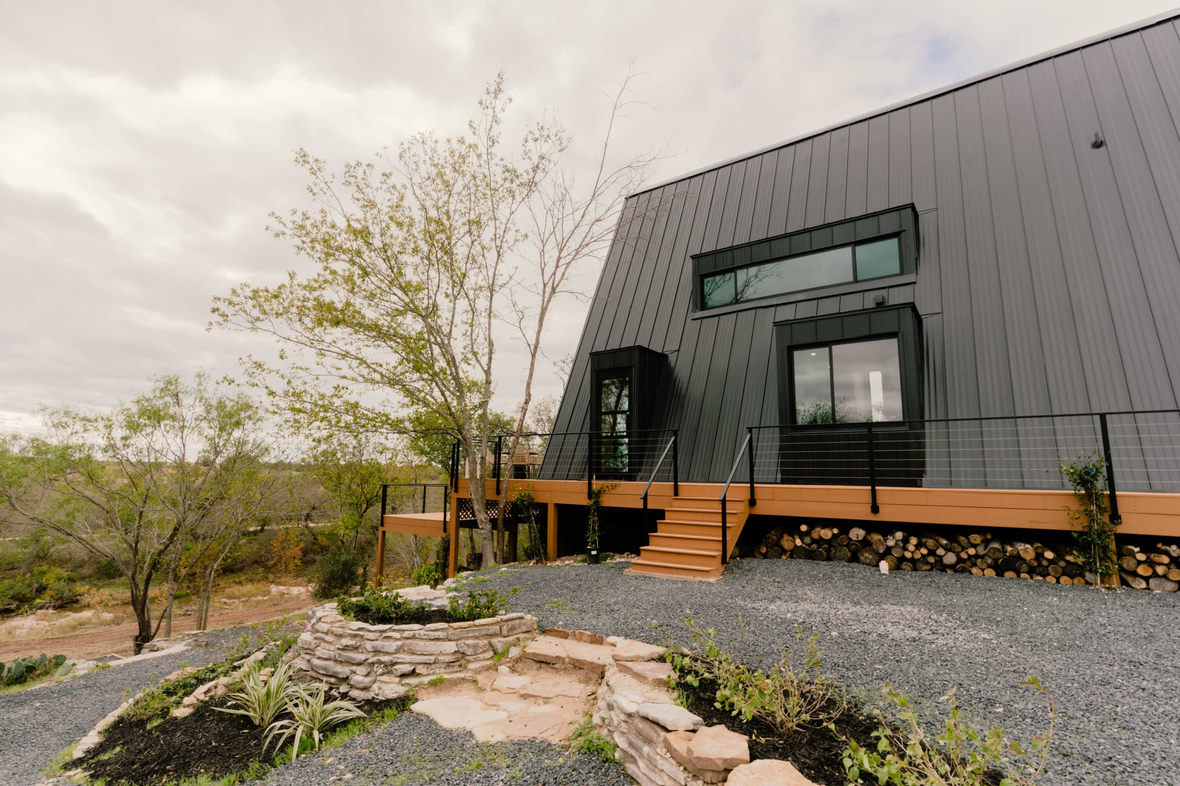 A modern, angular black house features a wooden deck and is surrounded by landscaped stone paths and greenery.