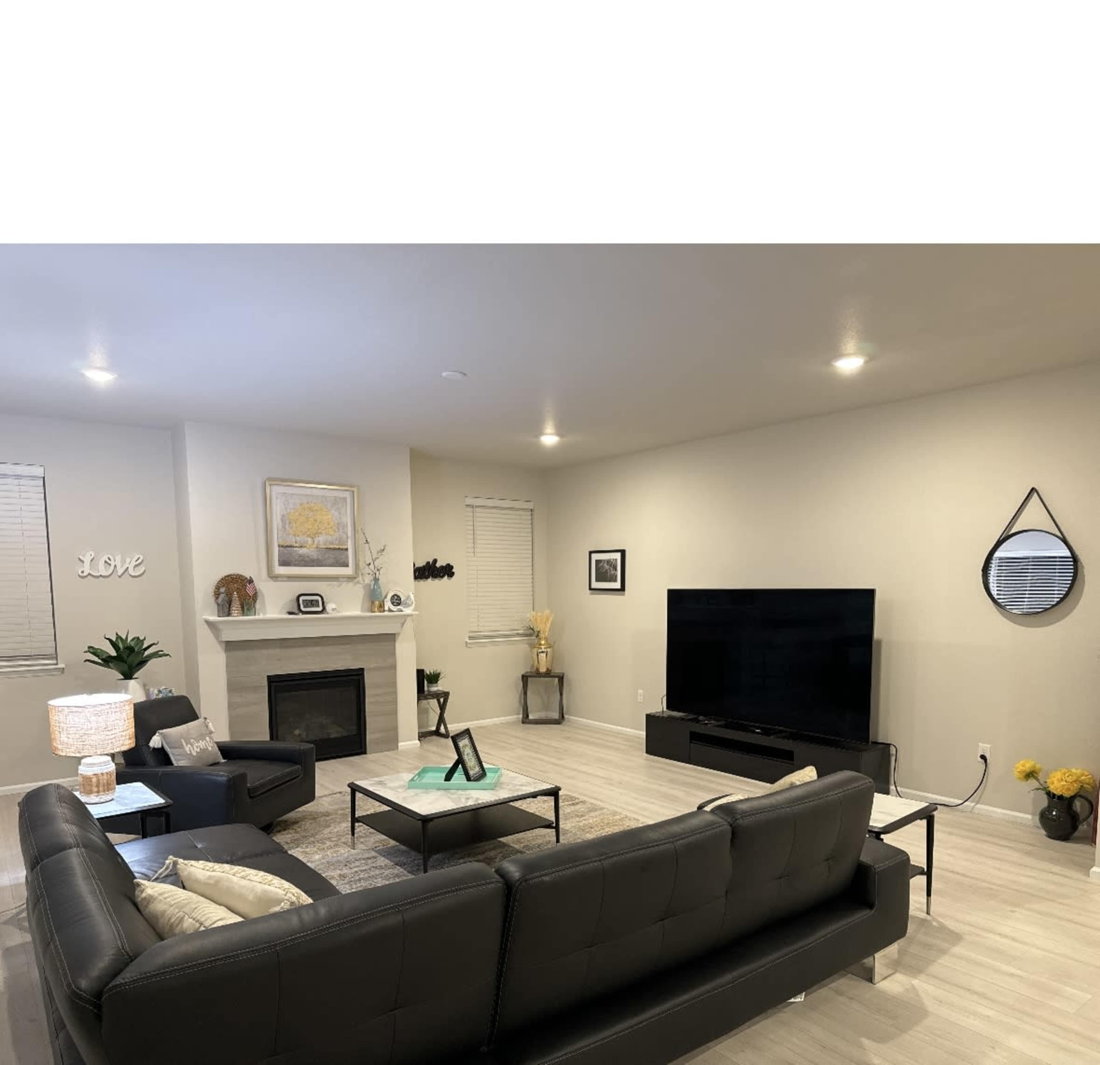 A modern living room features a black leather sofa set, a flat-screen TV on a media console, and a small fireplace, complemented by light-colored walls and wooden flooring.
