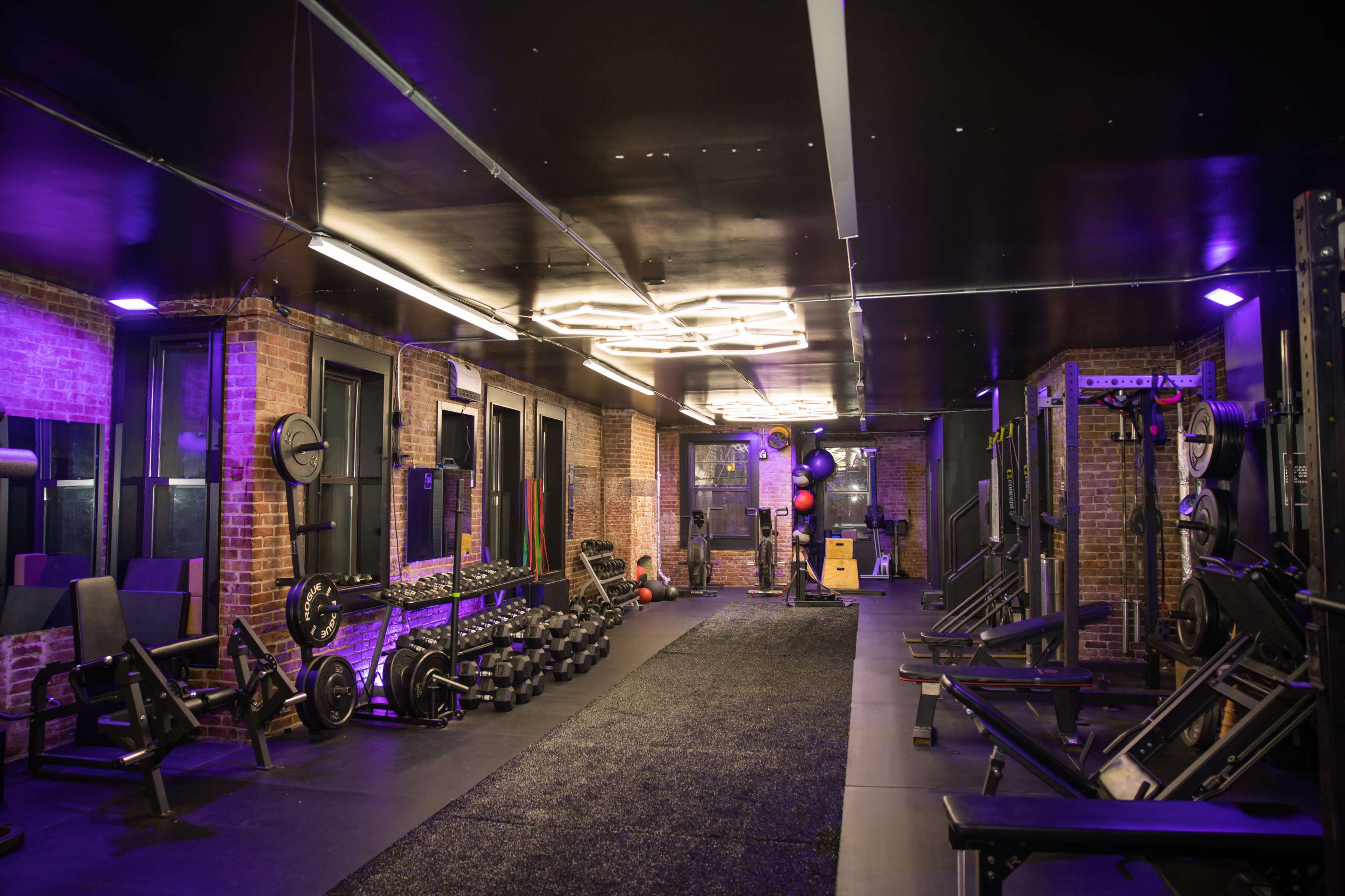 A gym interior features workout equipment, including weights and benches, with illuminated ceilings and exposed brick walls.
