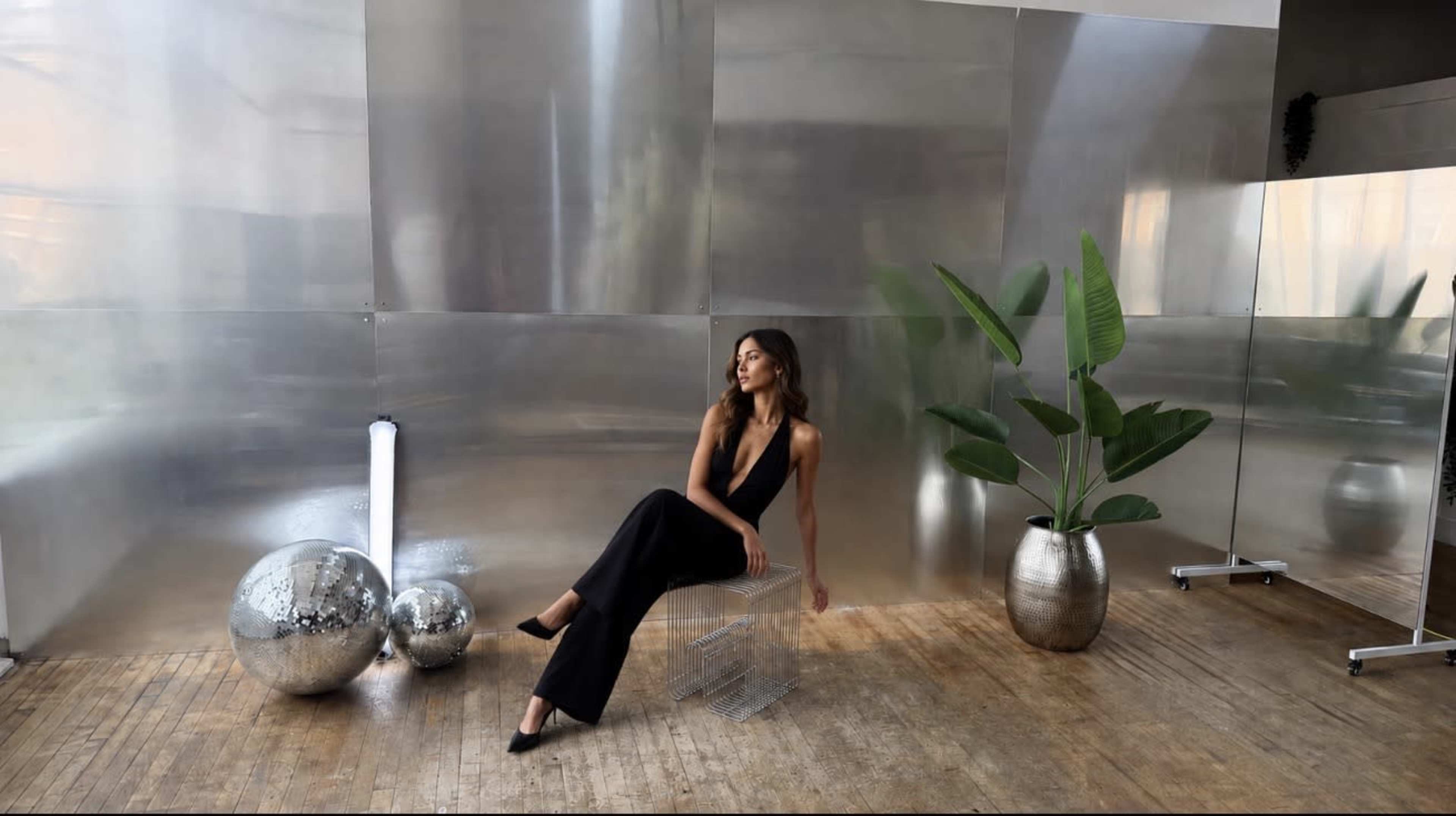 A woman in a black jumpsuit sits on a clear acrylic chair in a room with metallic walls, accompanied by two reflective spheres and a potted plant.
