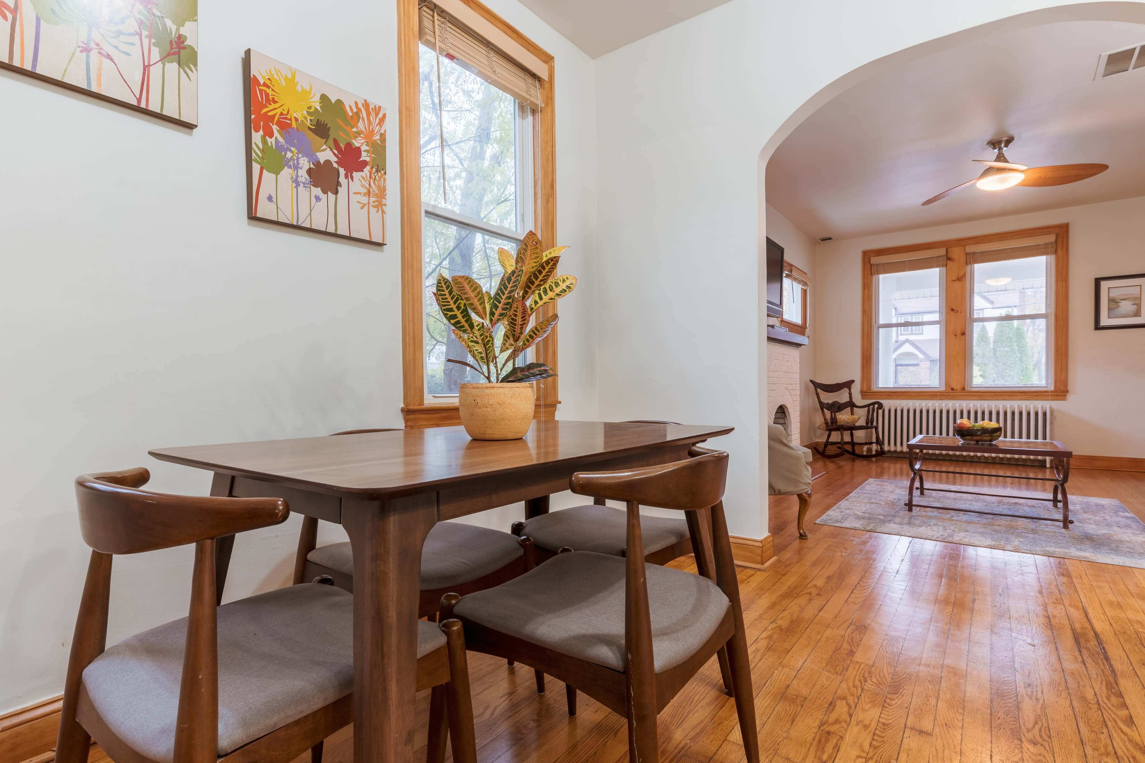 The image shows a dining area with a wooden table and chairs, adorned with a potted plant, next to a living room visible through an archway.