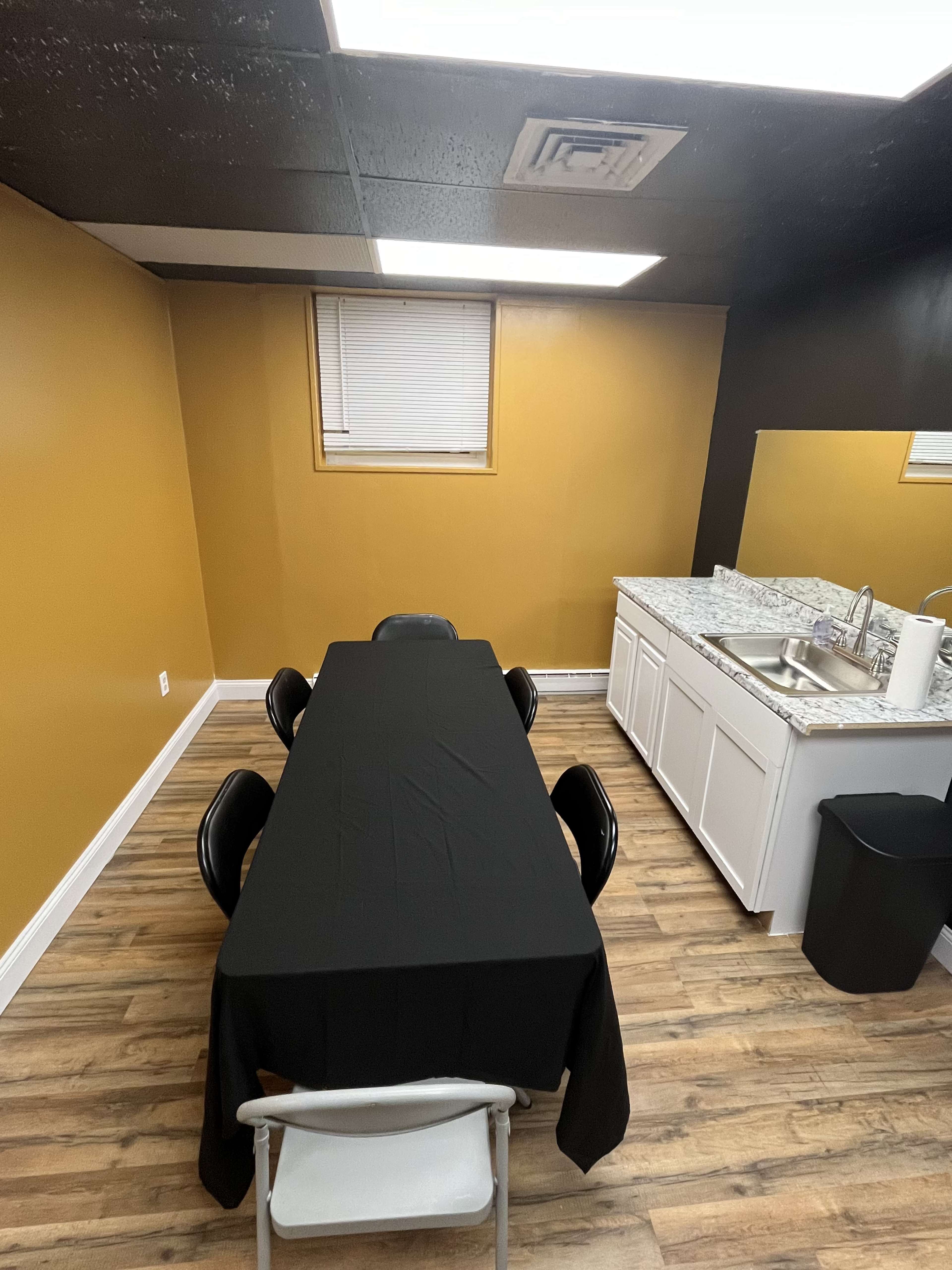 The image shows a small kitchen area with a black table set for six, surrounded by black chairs, next to a white countertop with a sink and a trash can.