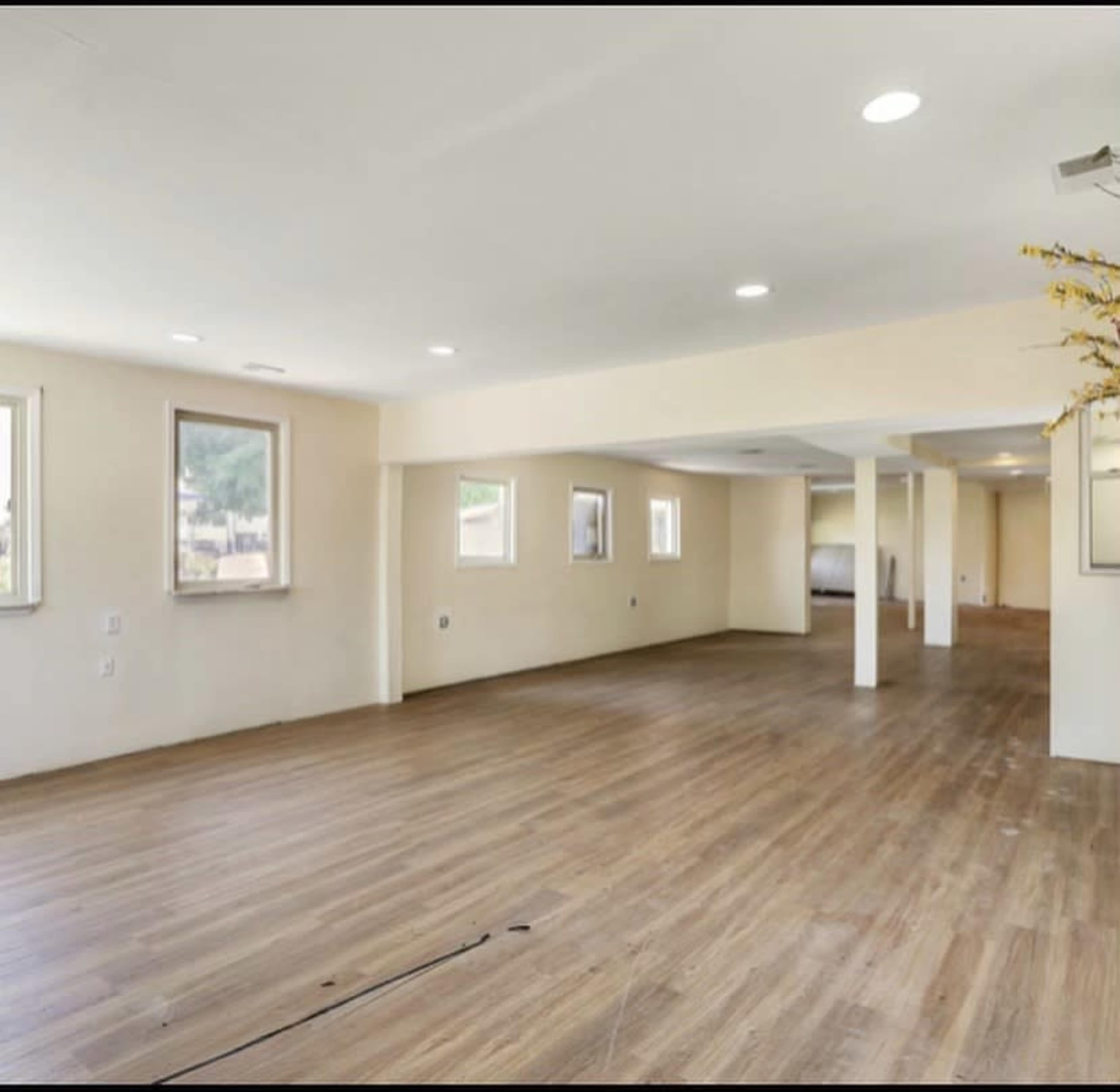 The image shows a spacious, empty room with wooden flooring and several windows allowing natural light.