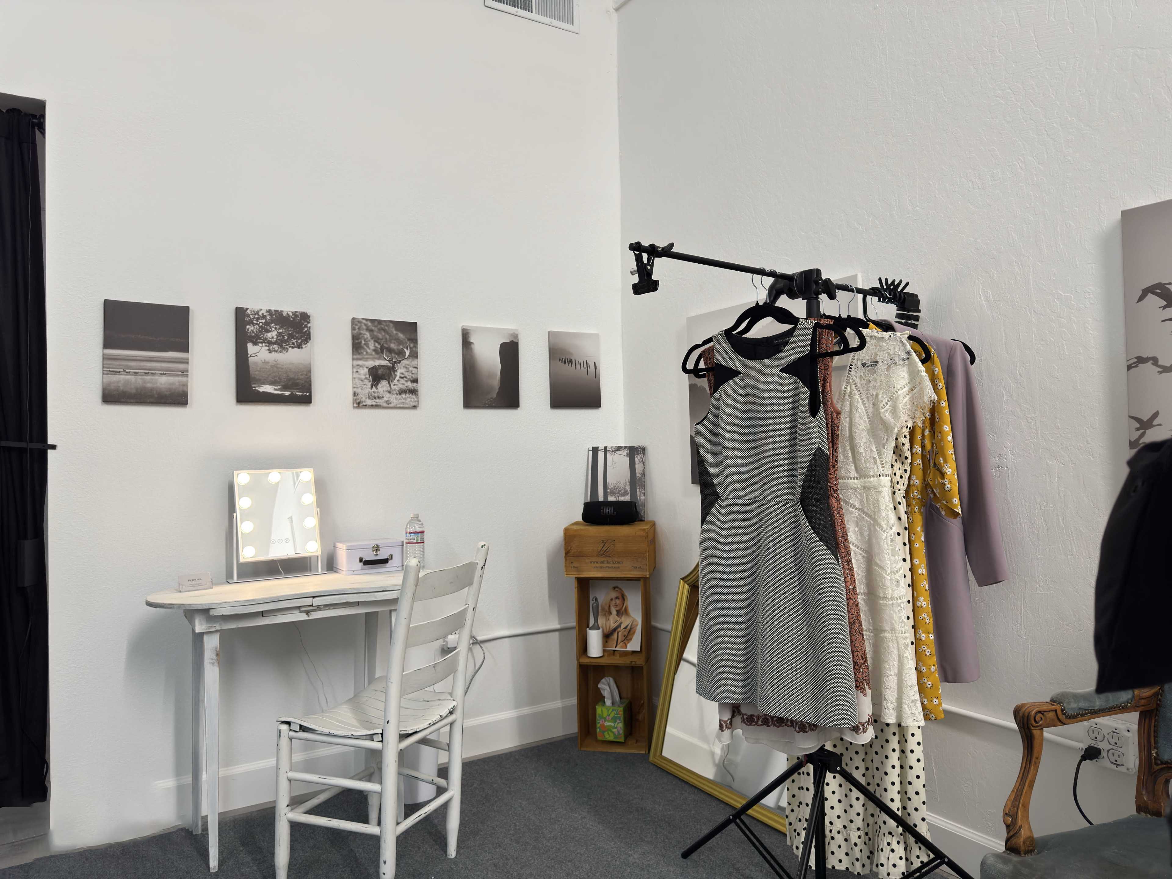 The image shows a small room with a clothing rack holding several dresses, a white desk with a lighted mirror, and black-and-white photographs mounted on the wall.