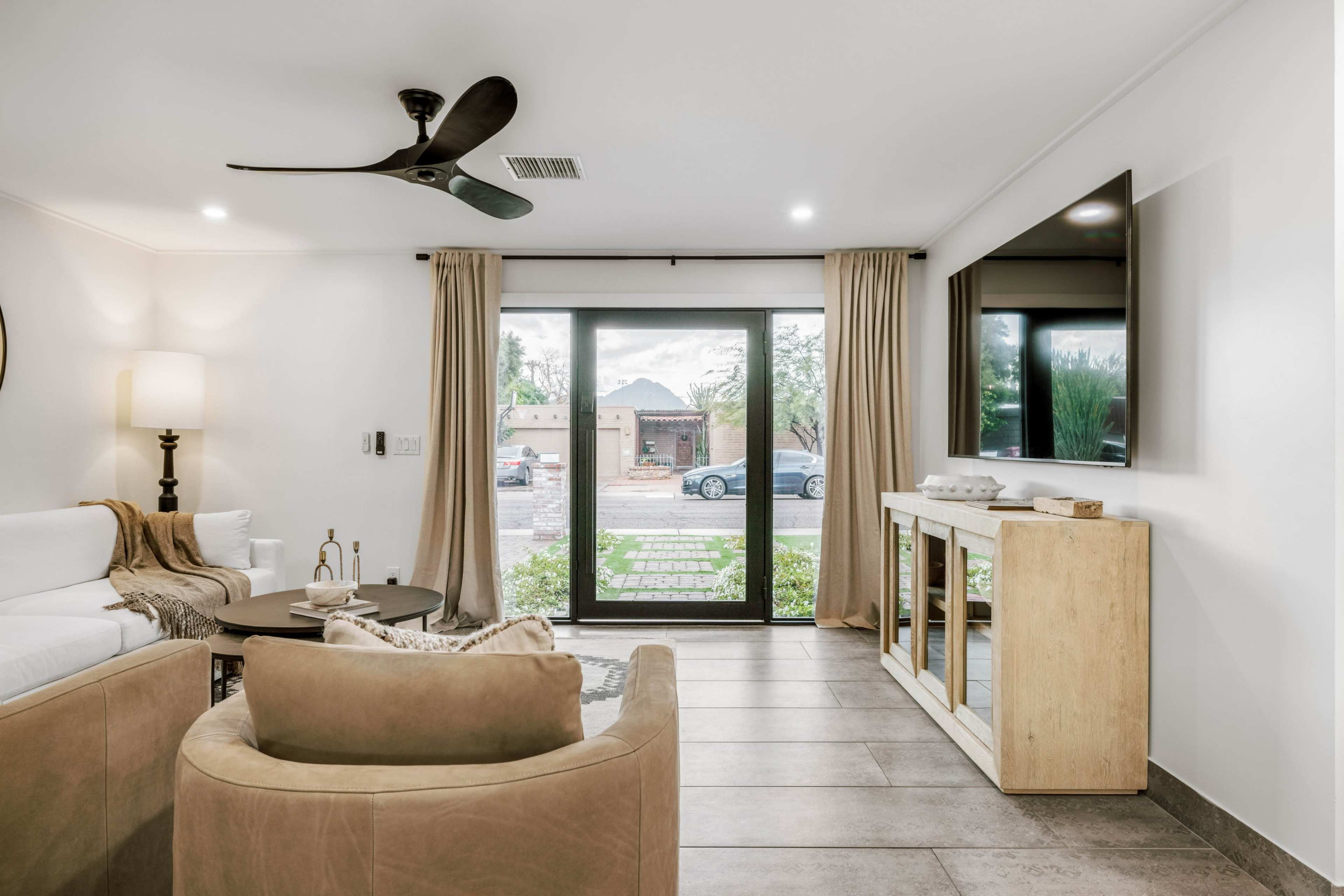 The image shows a modern living room with a beige sofa, a circular coffee table, and a television mounted on the wall, featuring large windows that overlook a landscaped exterior.