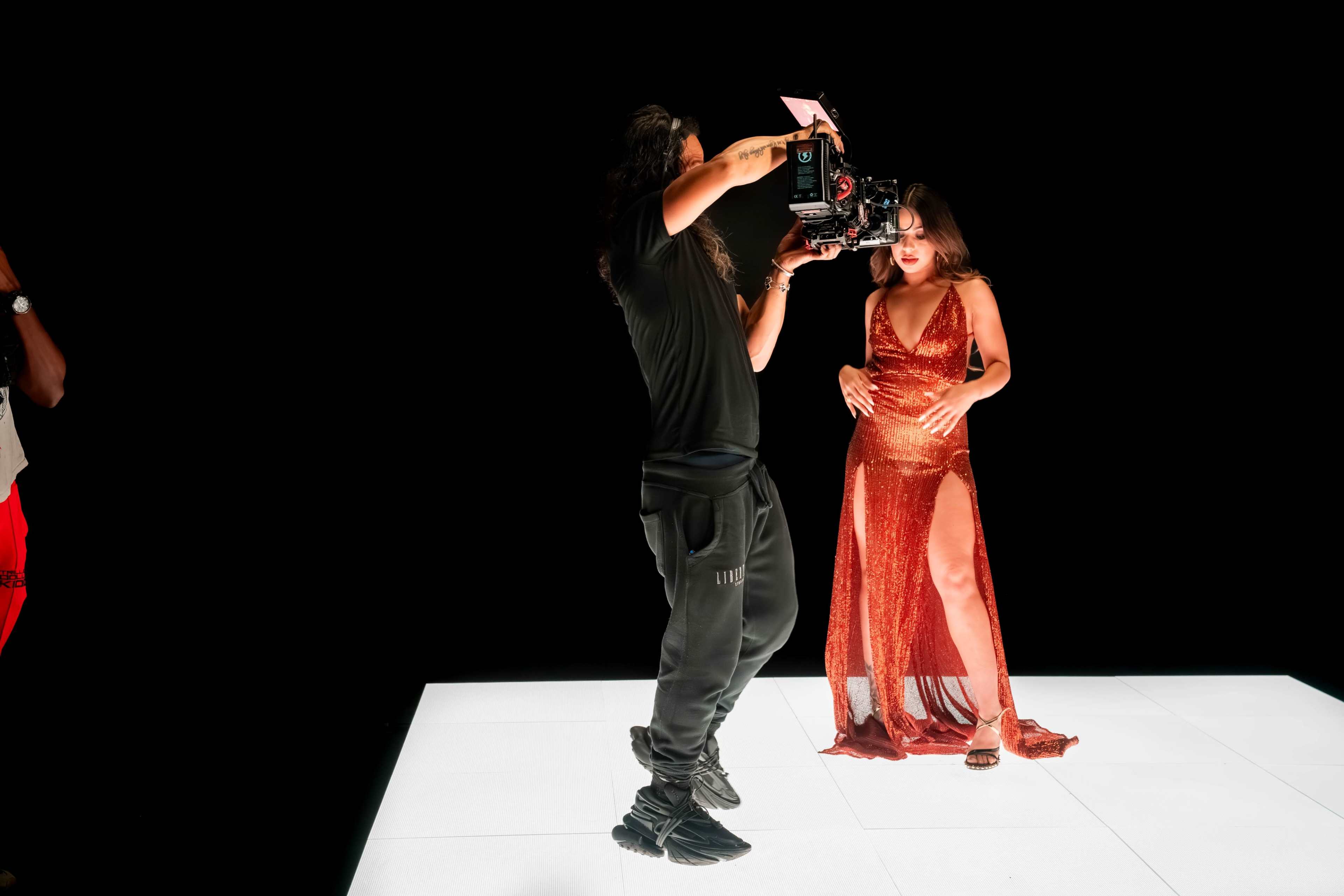 A person in a black shirt and pants operates a camera while a model in a shiny dress poses on a brightly lit platform against a black background.