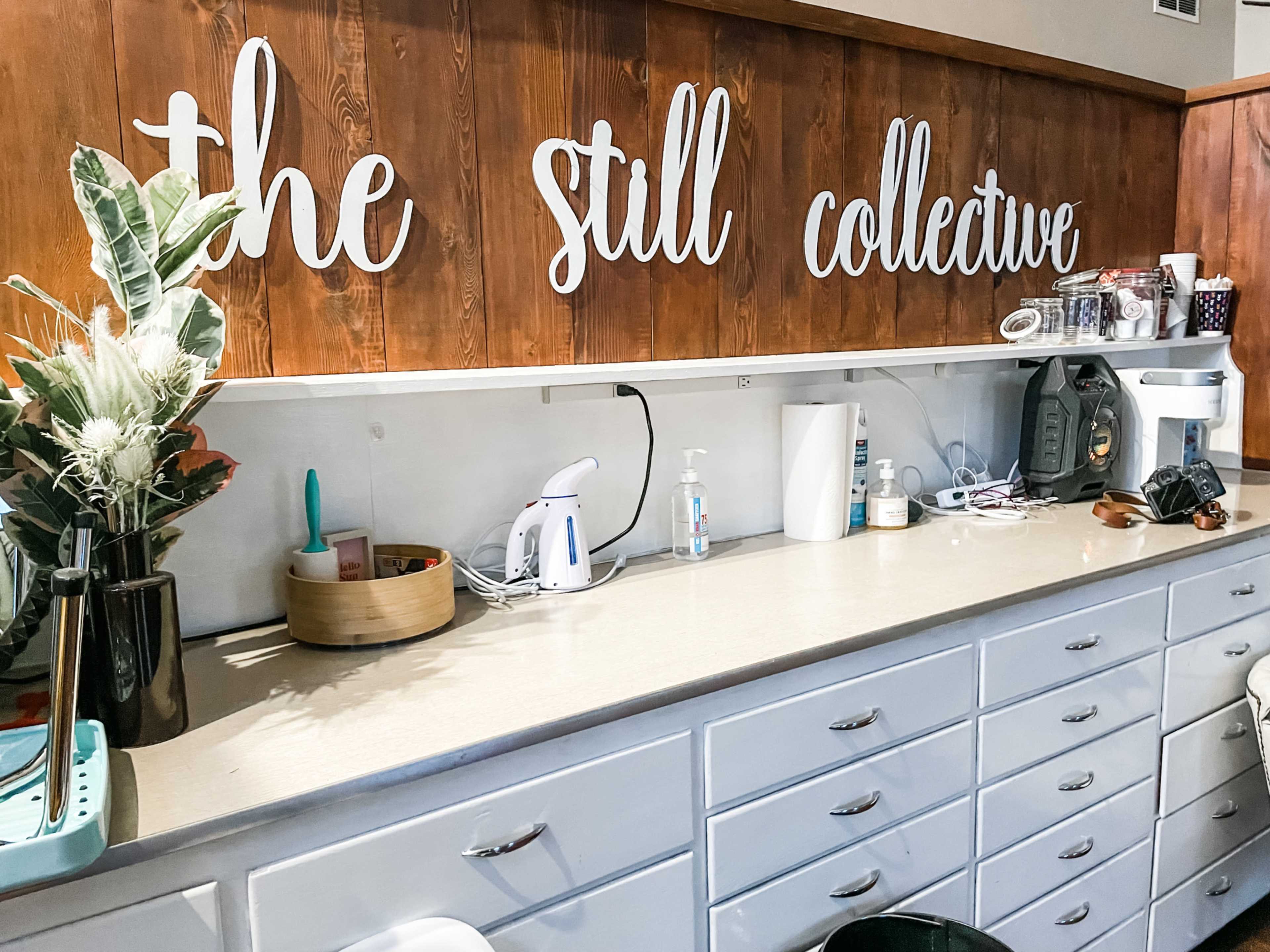 A countertop with various kitchen items, a plant in a black vase, and a sign that reads "the still collective" on a wooden wall.