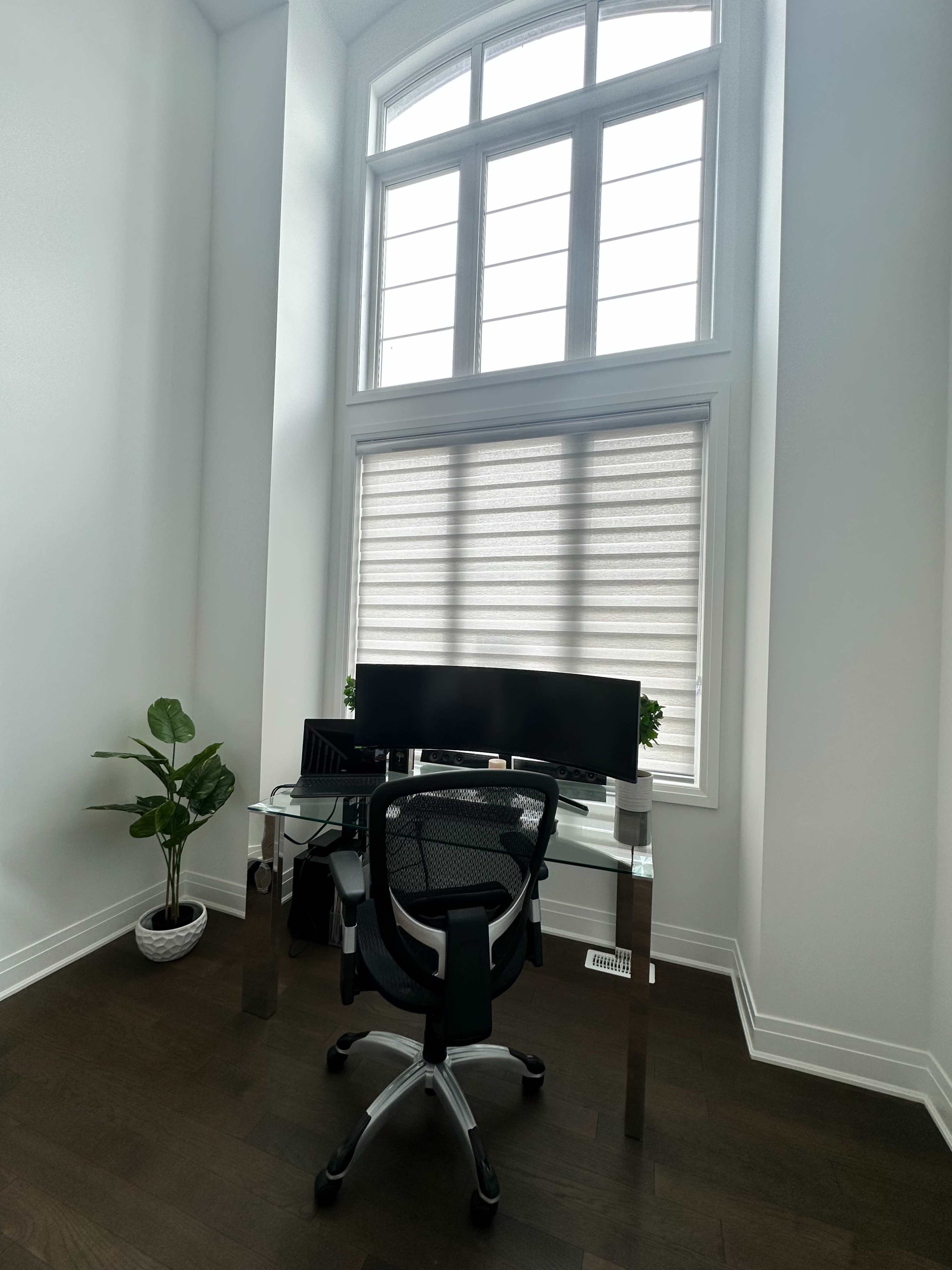 A minimalist home office features a glass desk with a computer monitor, an ergonomic chair, and a potted plant, illuminated by natural light from large windows.