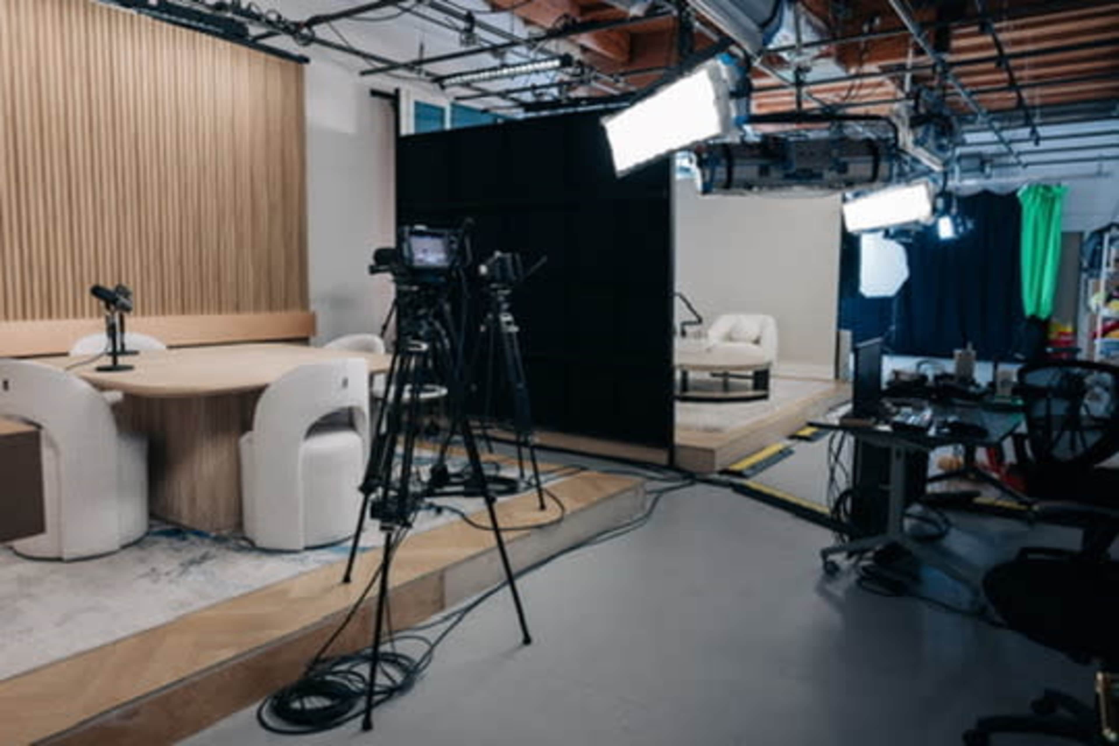 The image shows a television studio setup with multiple cameras, microphones, and a seating area, featuring a backdrop and various lighting equipment.