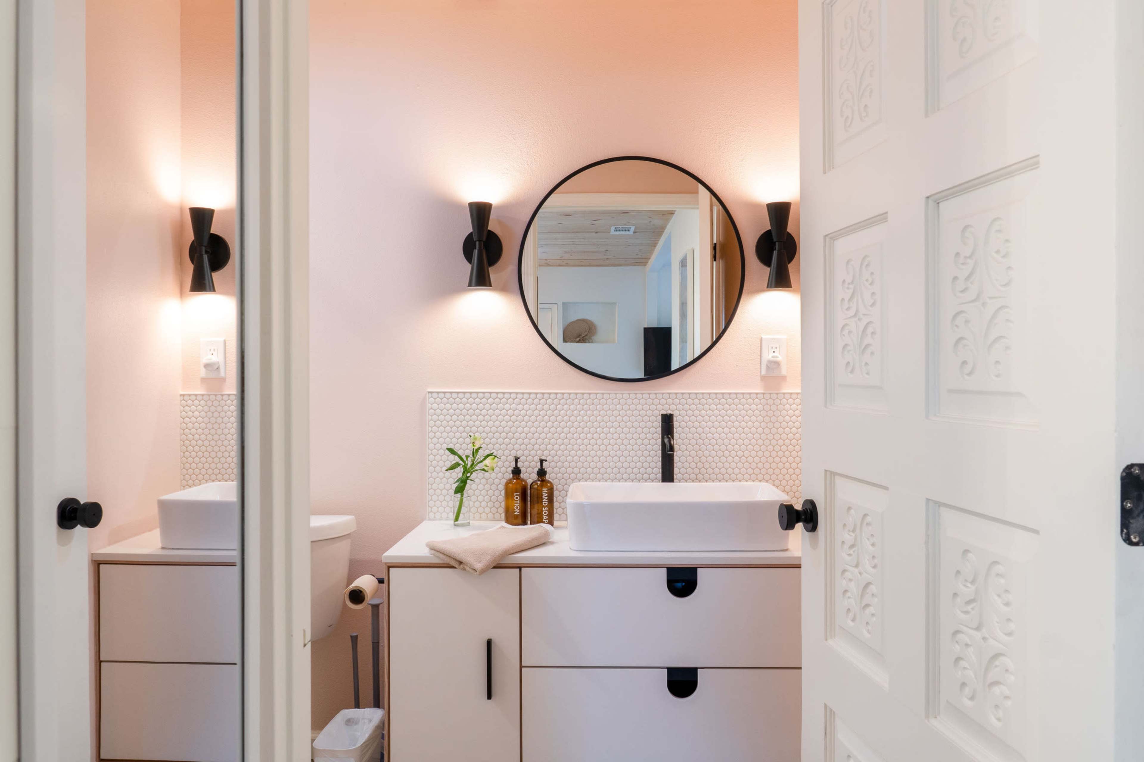 A modern bathroom features a circular mirror above a white sink, with black sconces illuminating the space against a light pink wall.