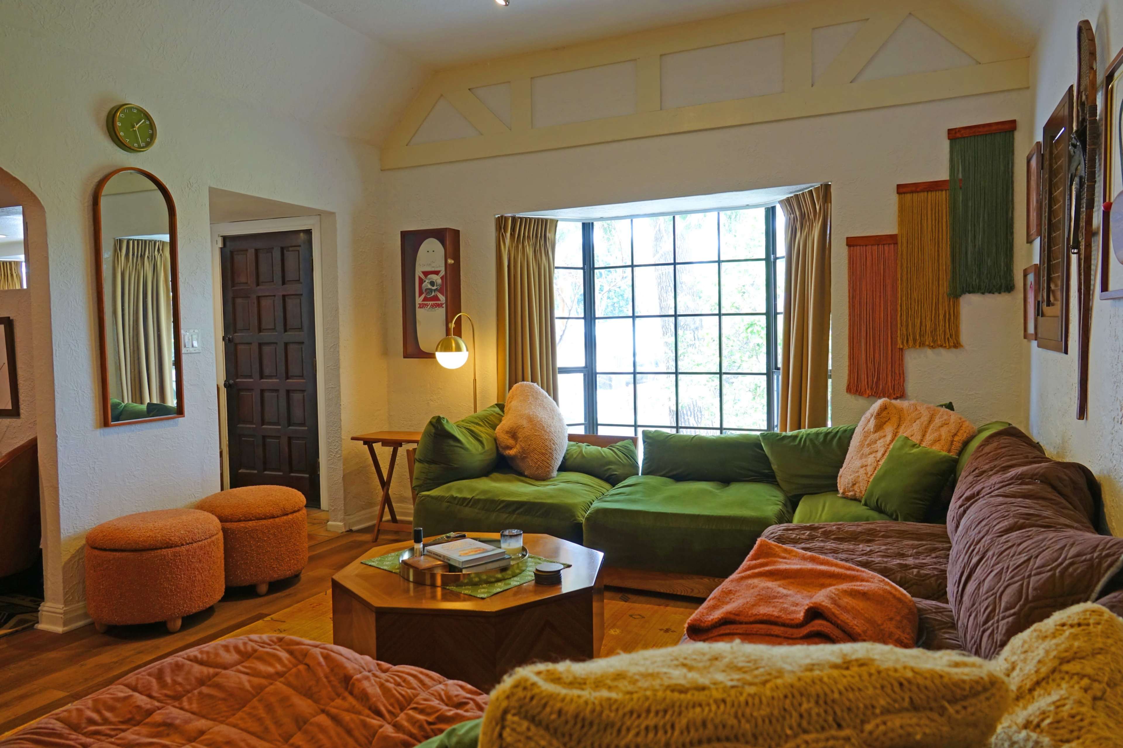 The image shows a cozy living room with green and orange furniture, a wooden table, and a large window letting in natural light.