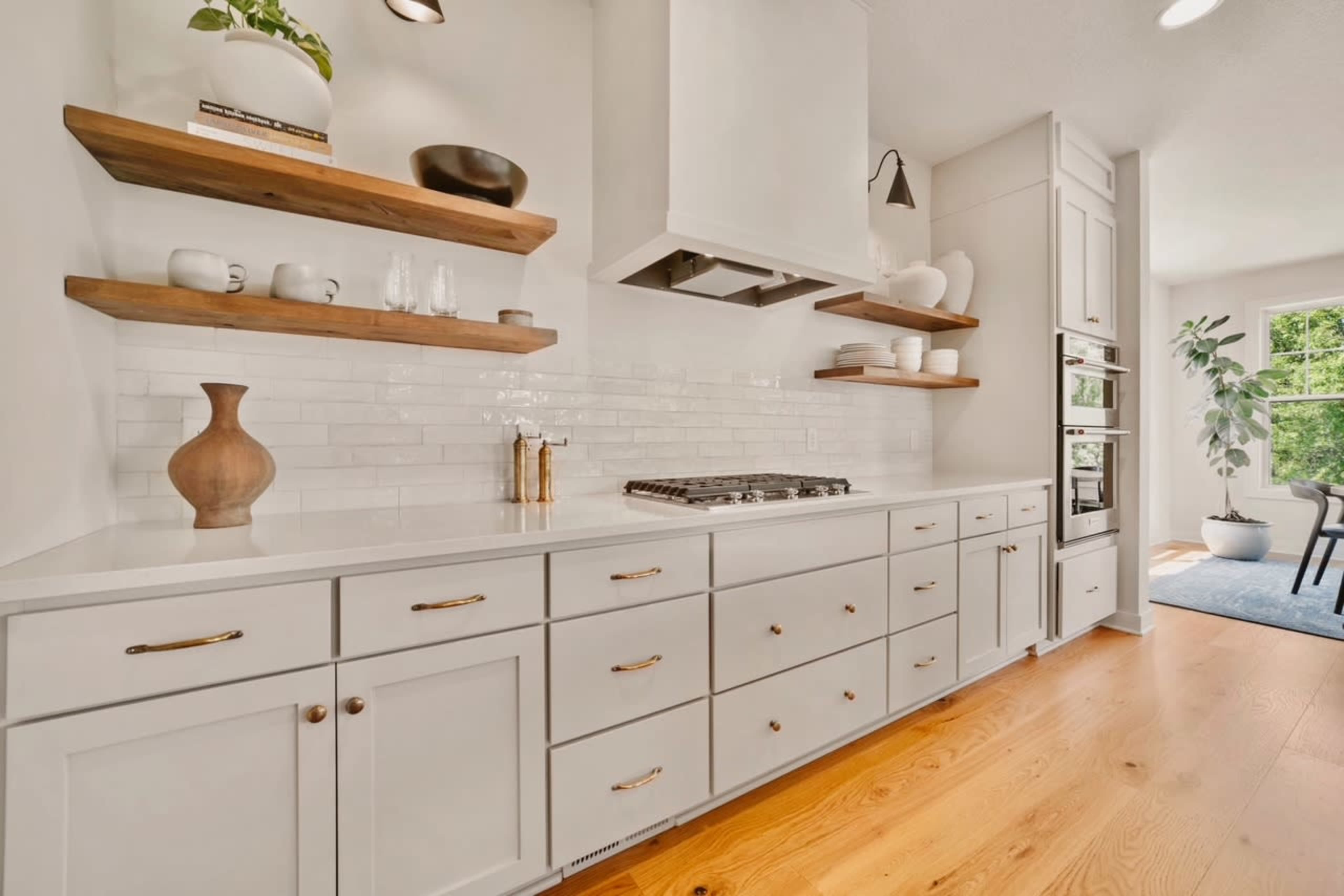 The image shows a modern kitchen featuring white cabinetry, wooden open shelves, and a gas stovetop beneath a white hood, with light flooring and a window in the background.