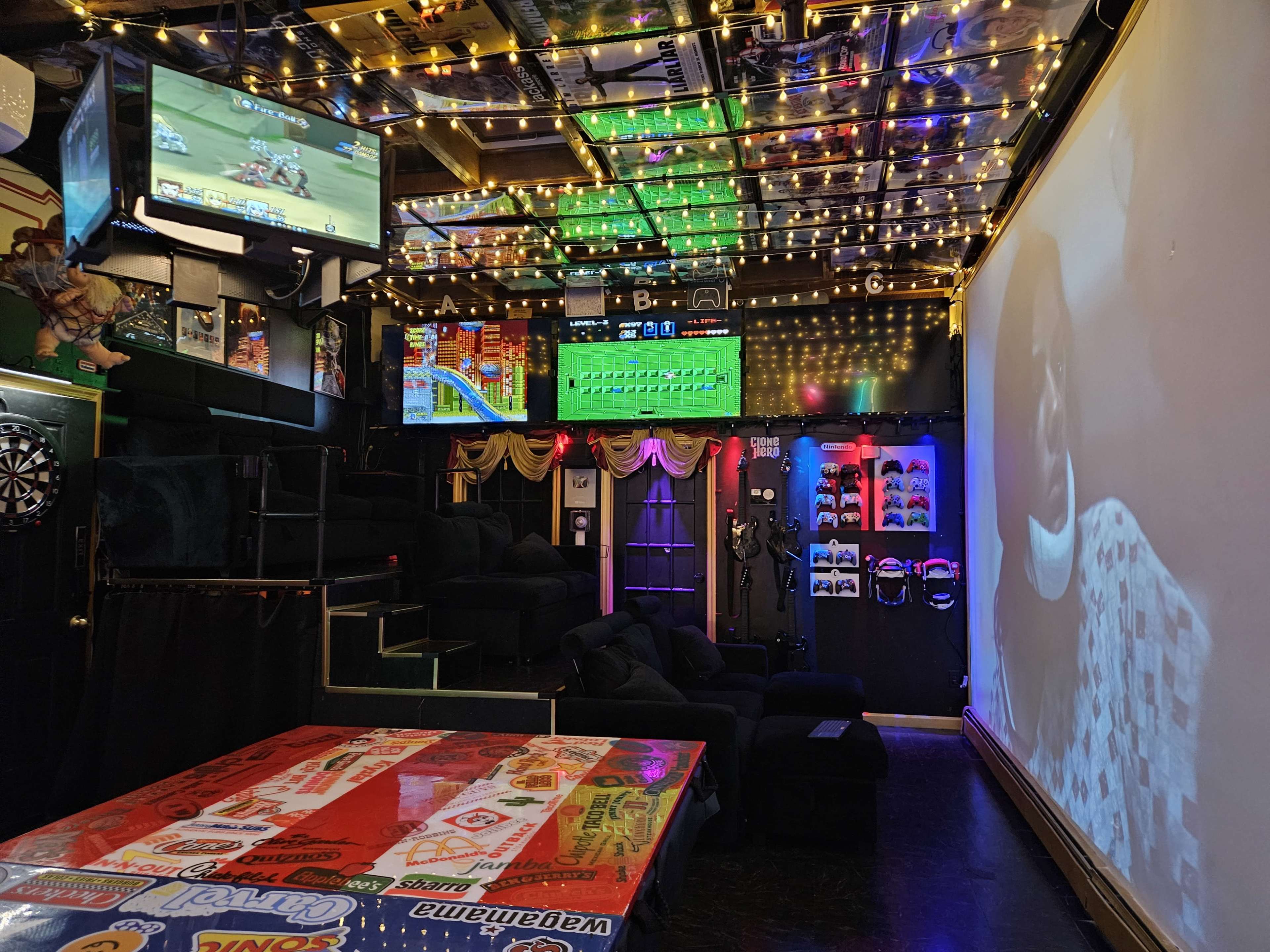 The image shows a dimly lit entertainment room featuring multiple screens displaying video games and a large projection on one wall, surrounded by colorful lights and decor.