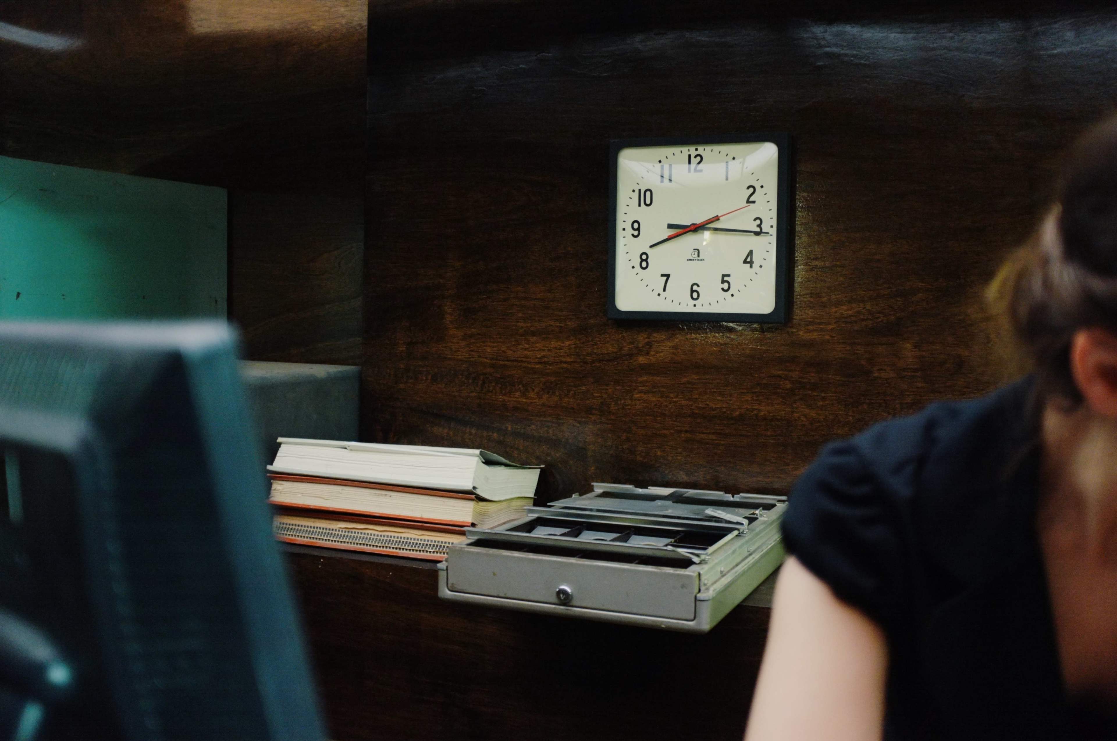 A wall clock showing 2:45 is mounted above a desk with a computer and a stack of books.