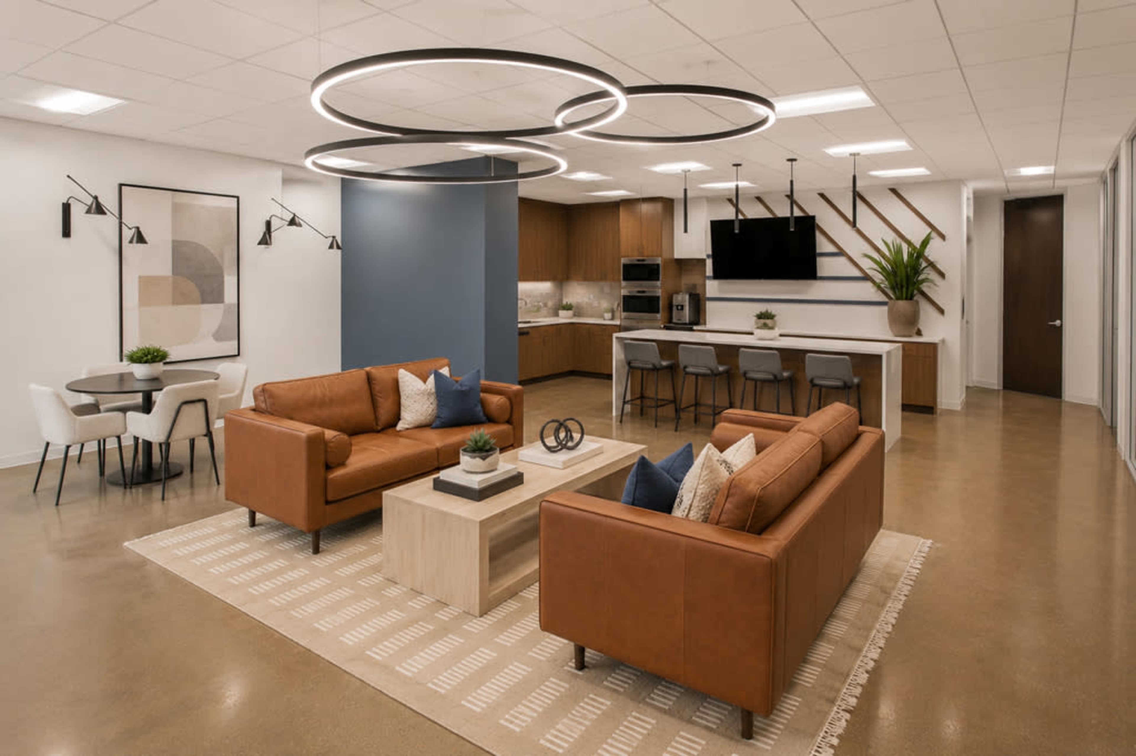 The image shows a modern lounge area featuring two brown leather sofas, a coffee table, and a kitchen space with bar seating in the background.