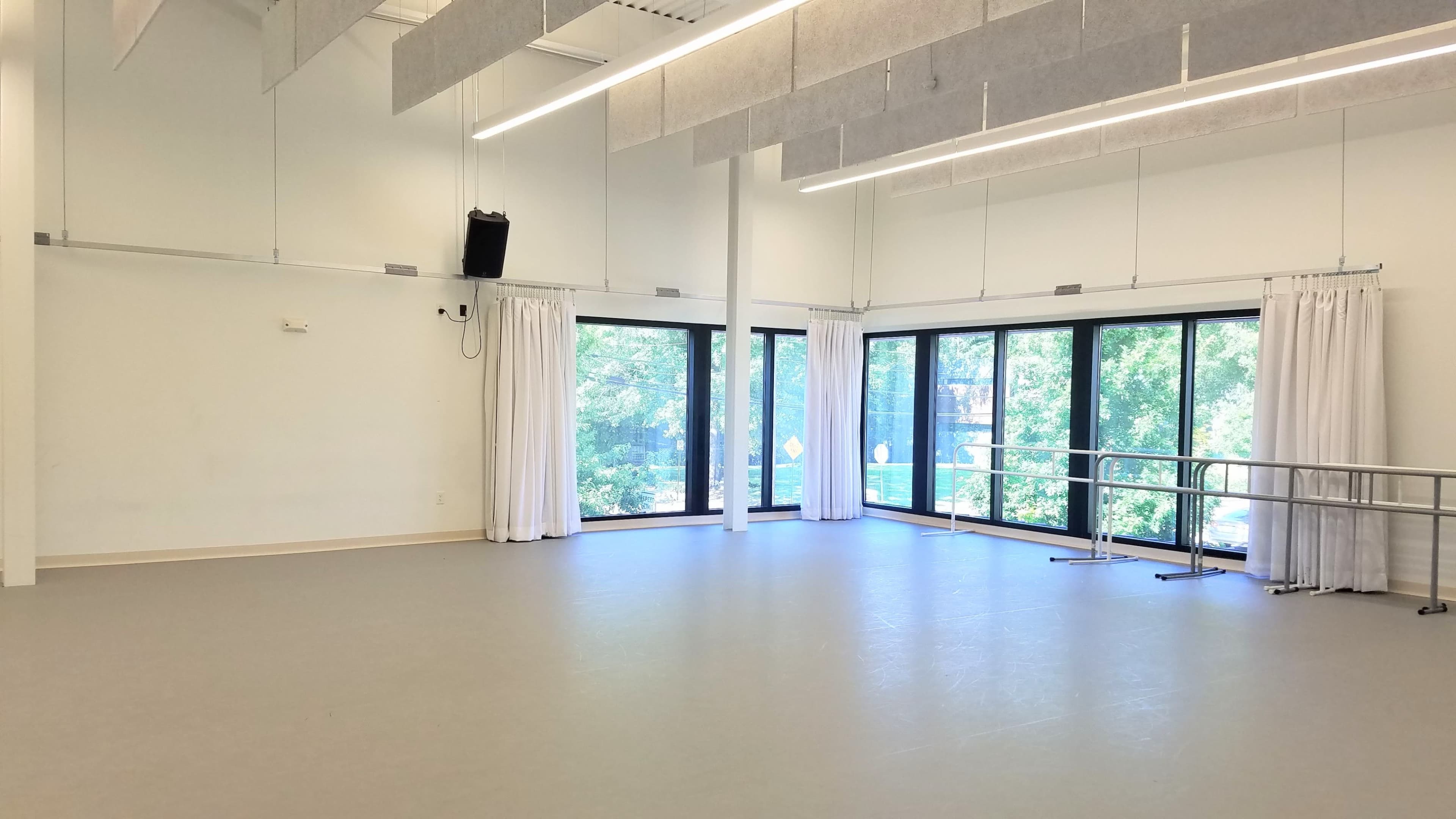 The image shows a spacious, well-lit room with large windows, light-colored walls, and a ballet barre along one side.