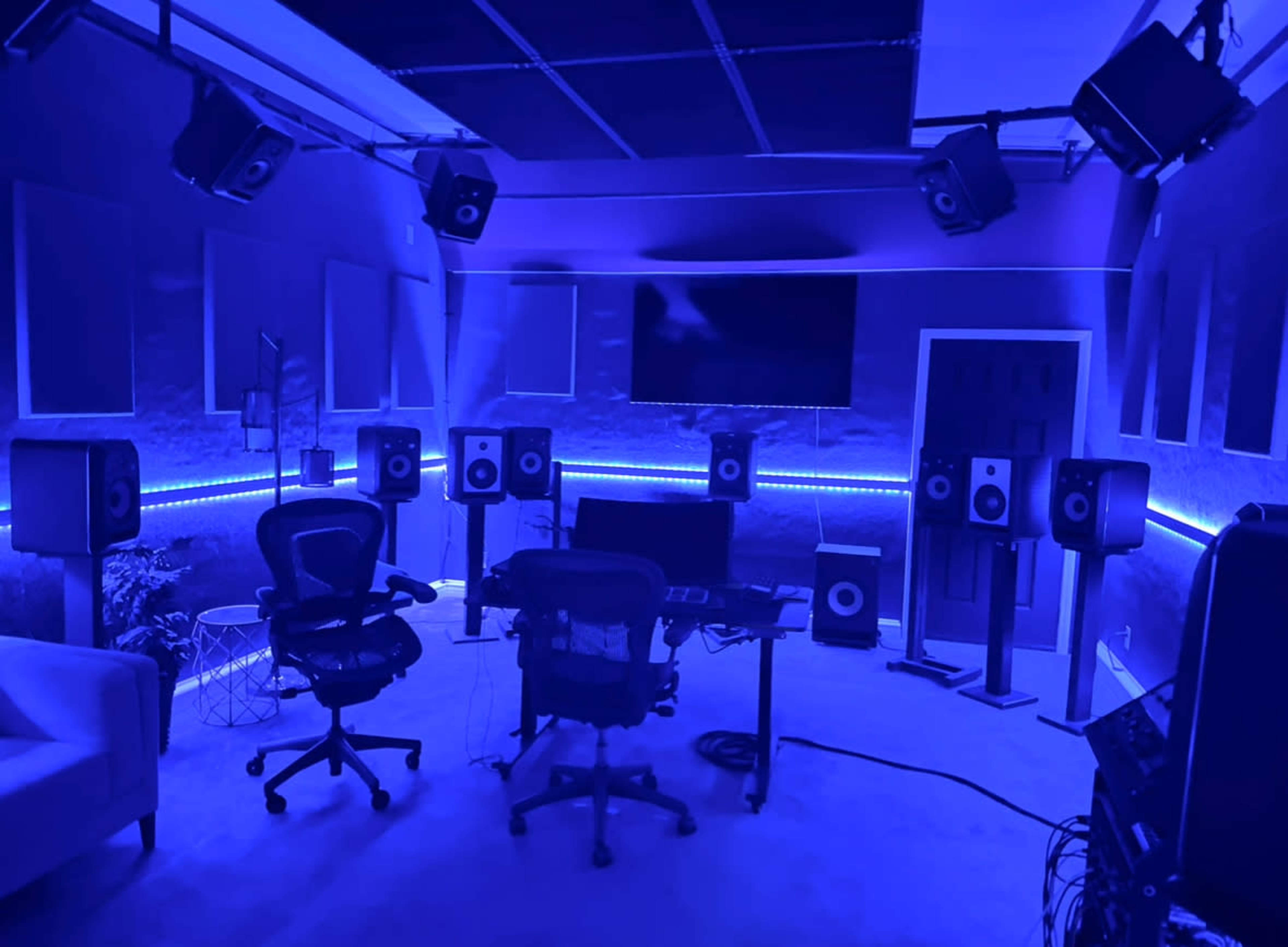 A music studio features multiple speakers, a central desk with chairs, and a large screen illuminated by blue lighting.