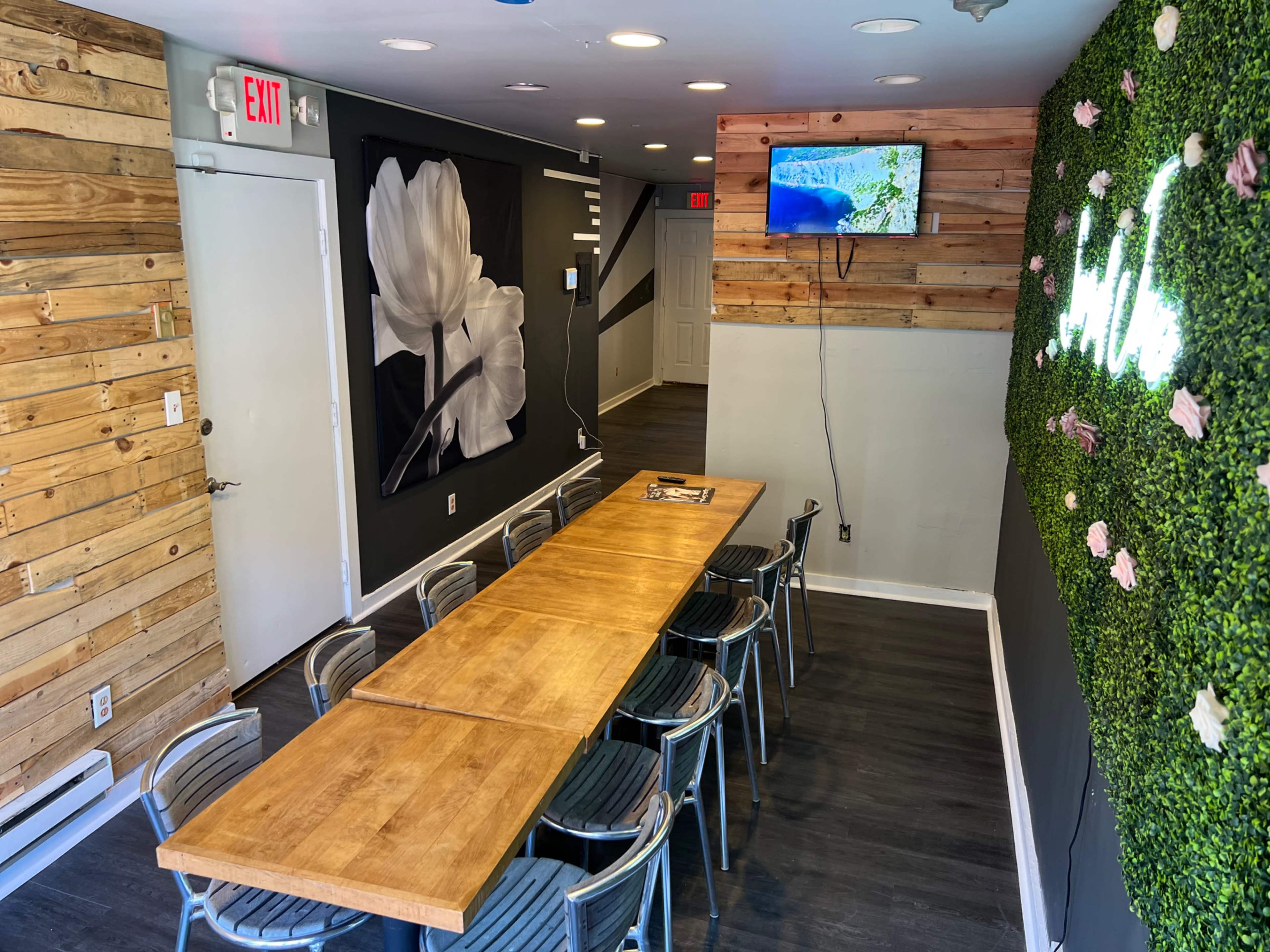 A dining area with a long wooden table surrounded by metal chairs, a television mounted on the wall, and a green moss wall feature.