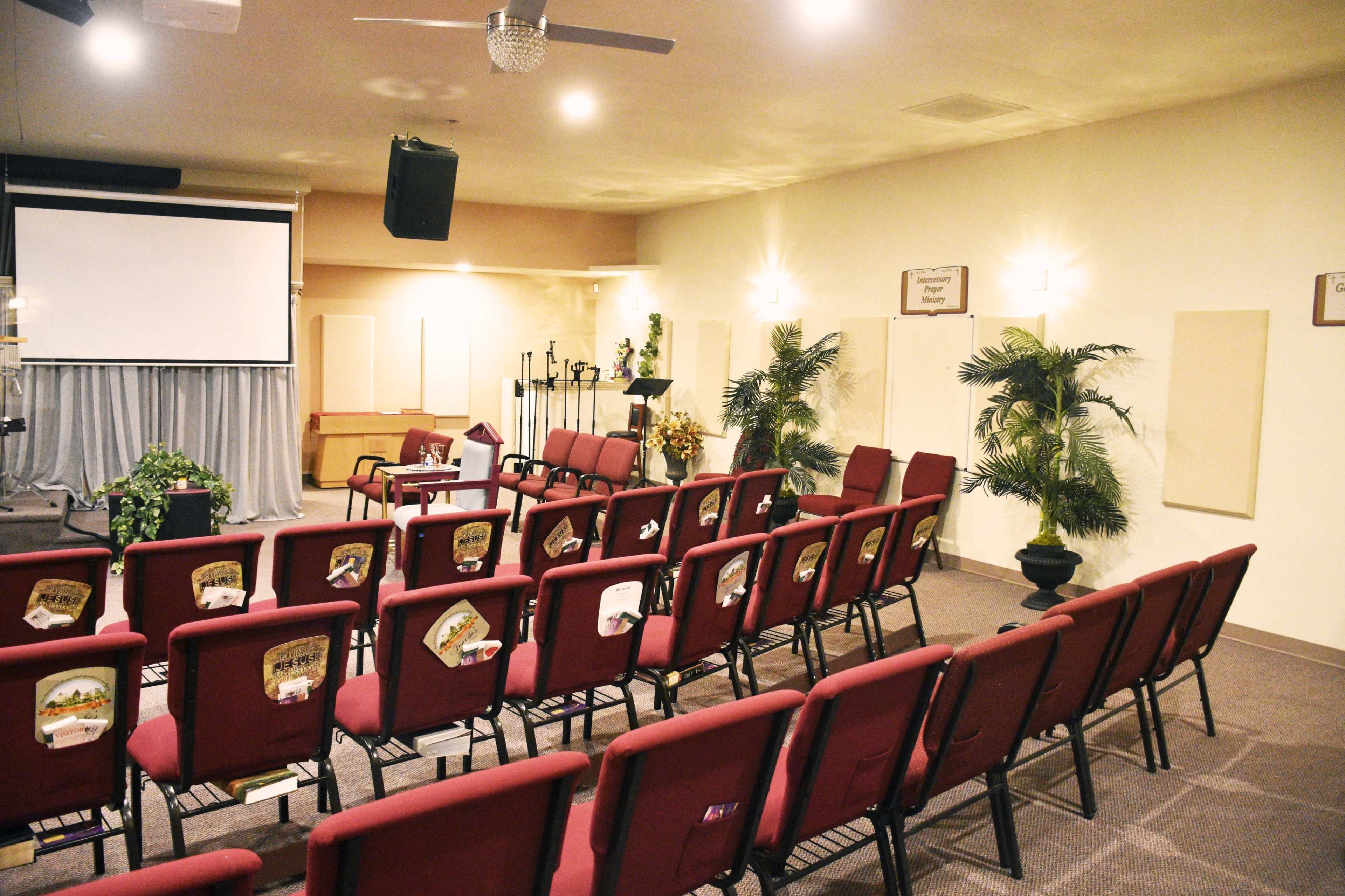 An empty church interior with rows of burgundy chairs facing a stage area and decorative plants in the corners.
