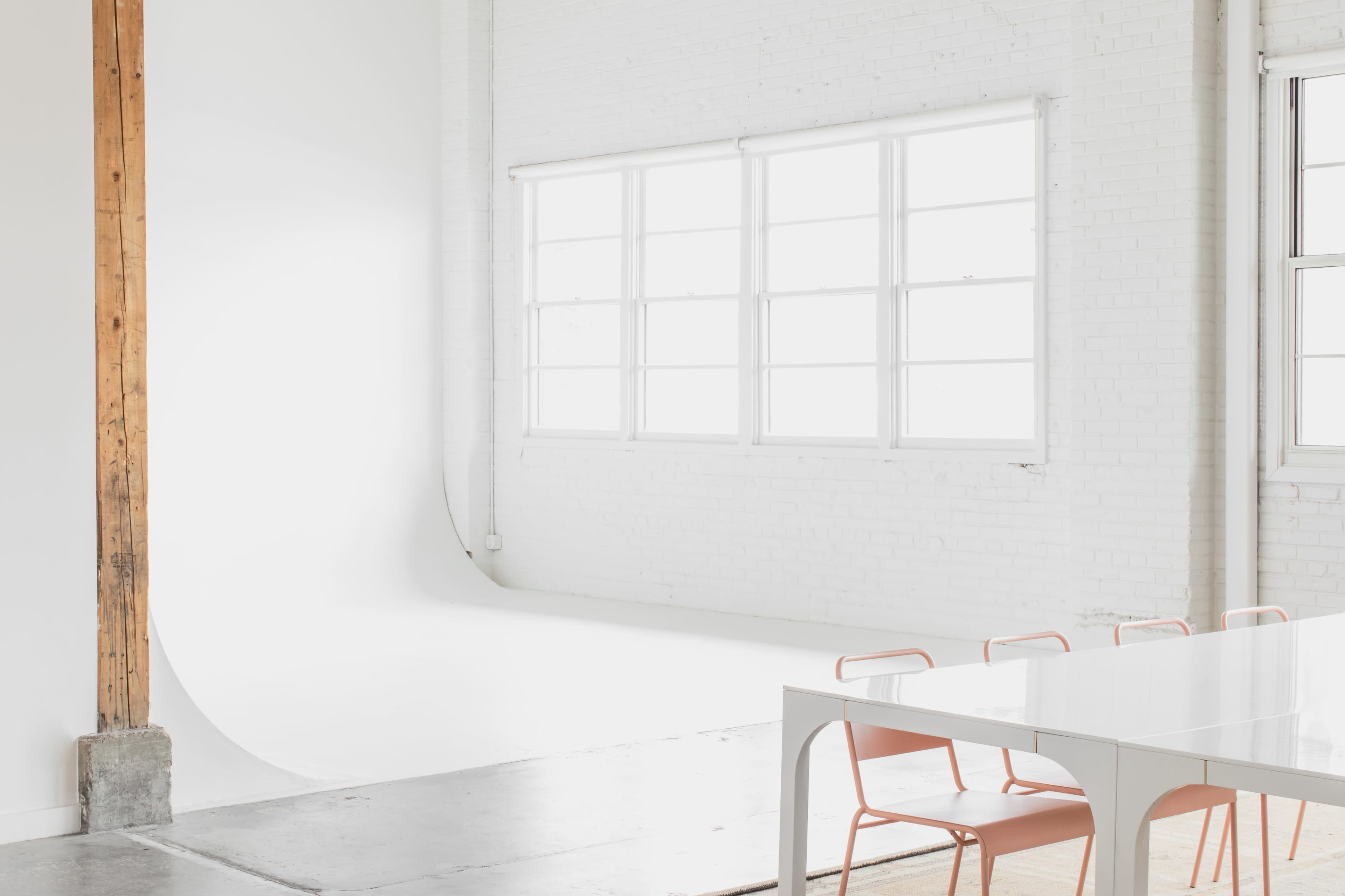 The image shows a minimalist photography studio with large windows, white walls, and a simple table with pink chairs.