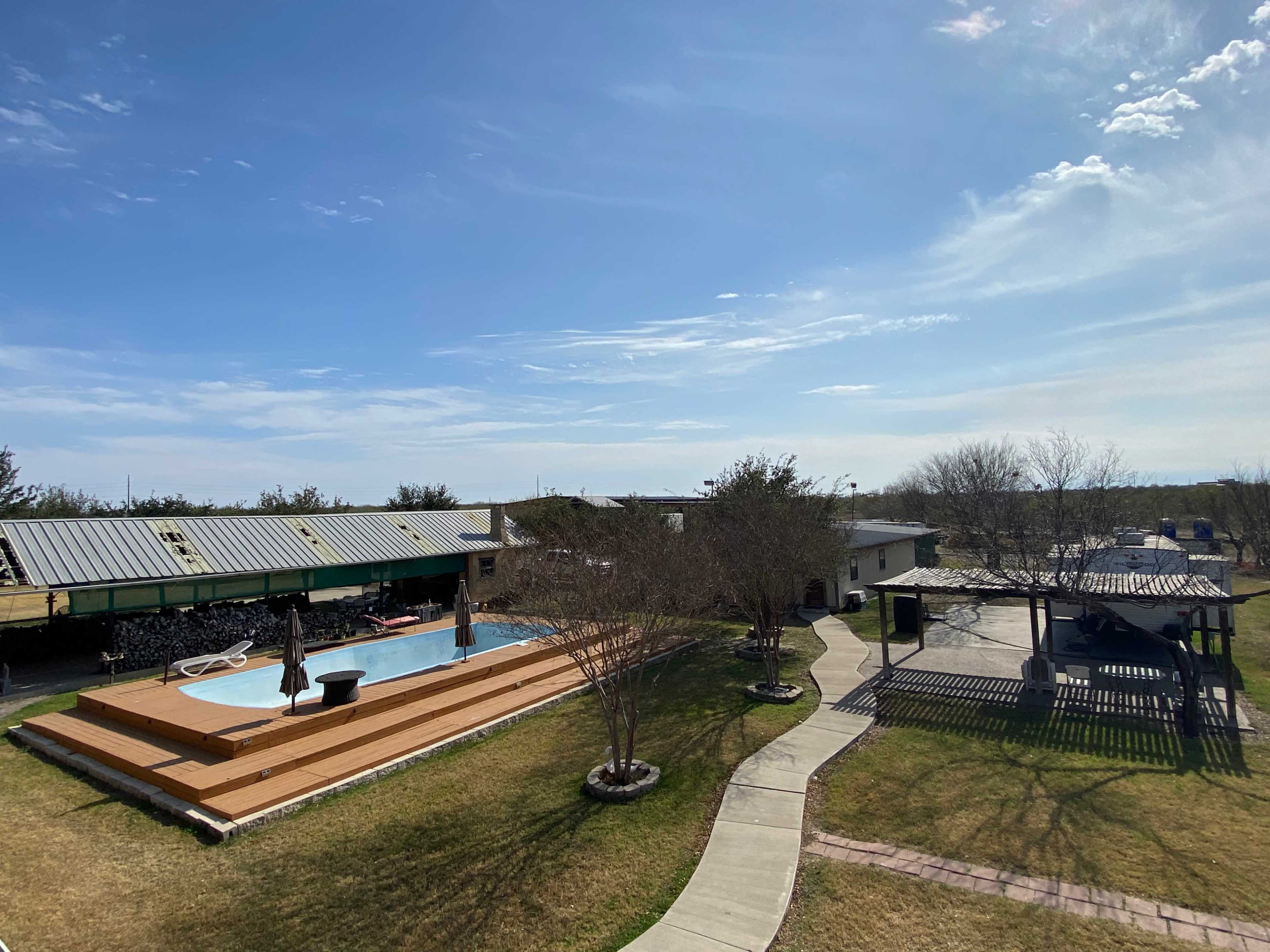 The image shows a backyard with a swimming pool surrounded by wooden decking, a paved pathway, and several buildings under a clear blue sky.