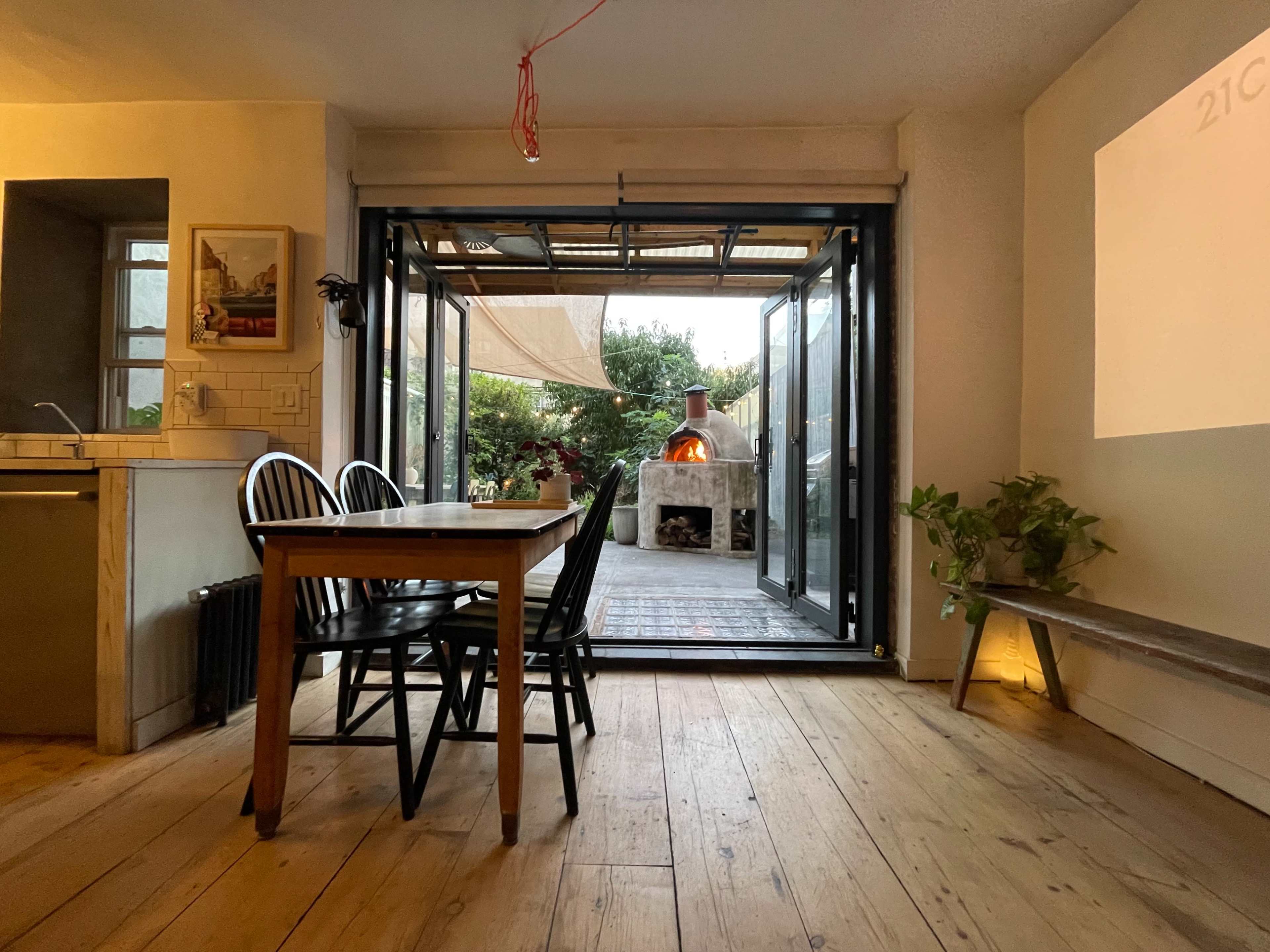 A dining area with a table and chairs opens to a patio featuring a wood-fired oven and lush greenery.