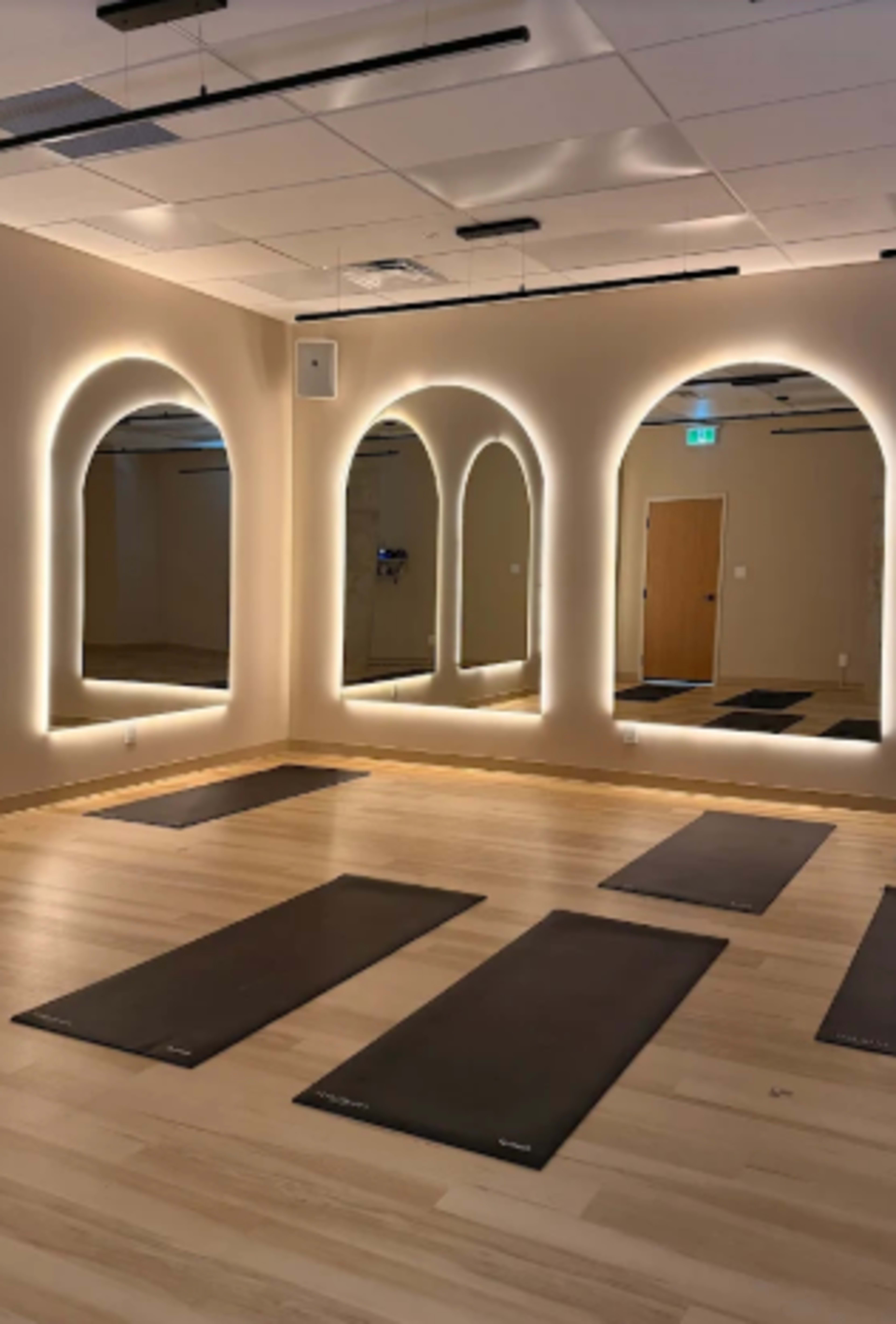 The image shows a yoga studio with several mats laid out on a wooden floor, illuminated by soft lighting and featuring large, arch-shaped mirrors on the walls.