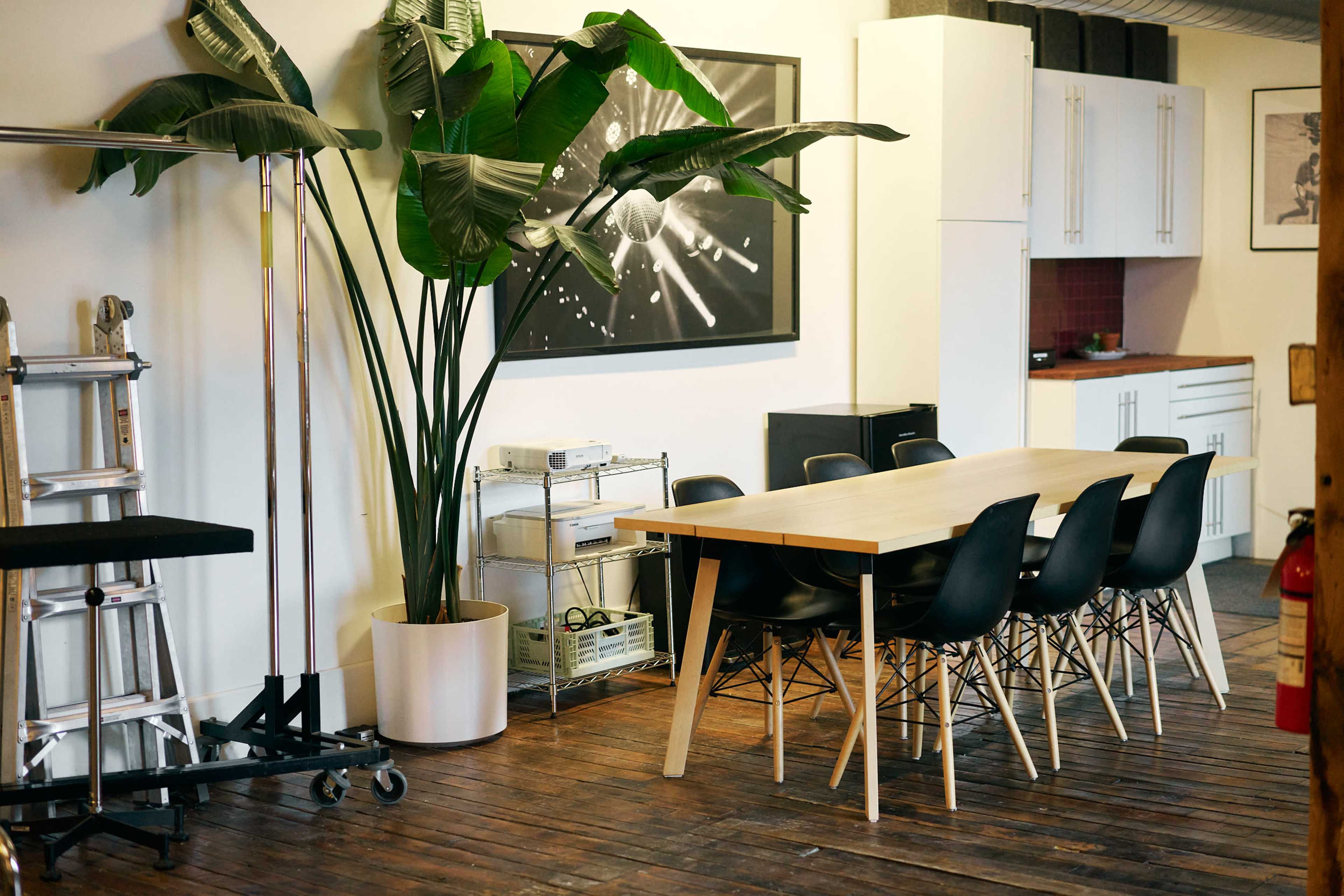 The image shows a modern workspace with a wooden dining table surrounded by black chairs, a tall potted plant, and a kitchen area in the background.