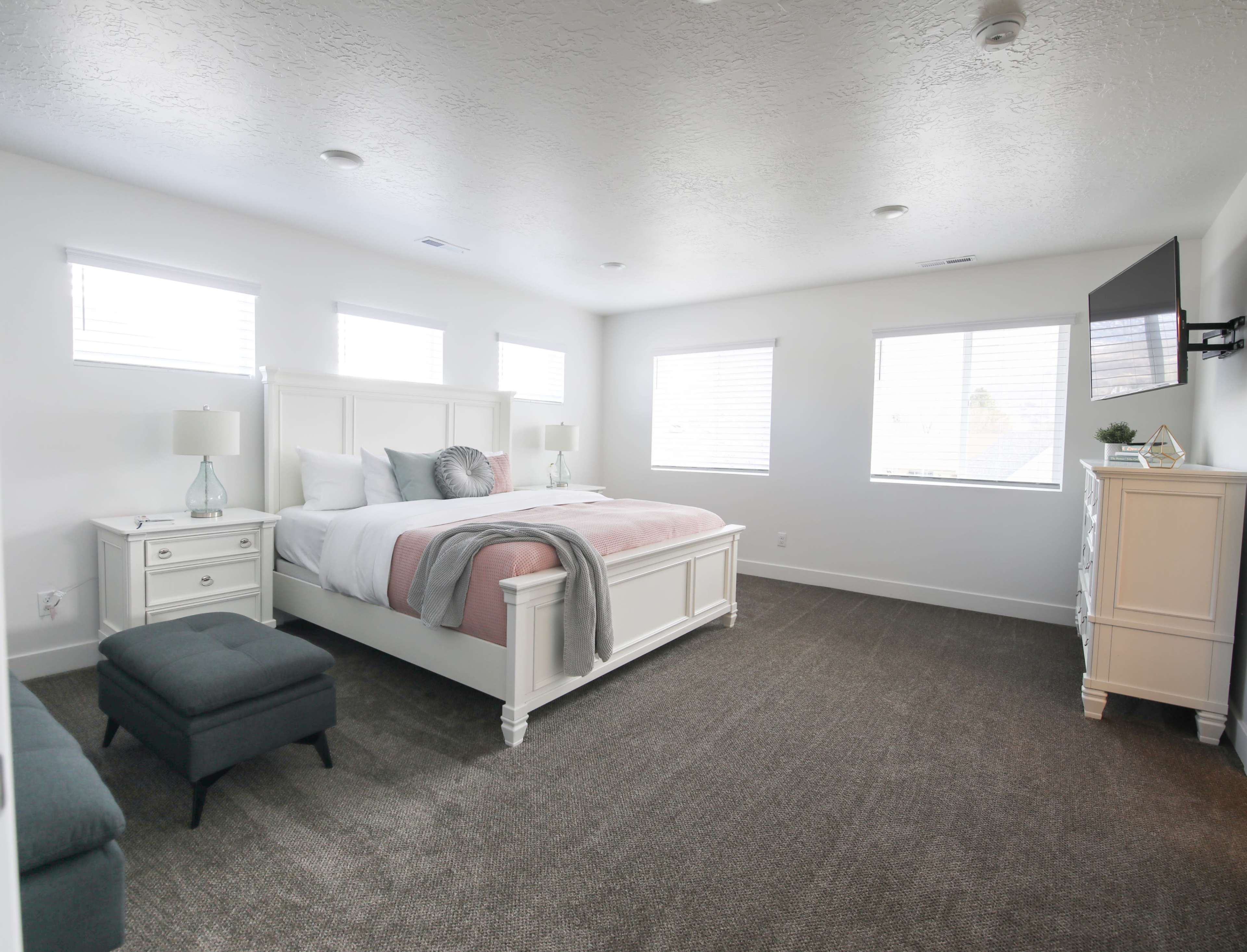 A well-lit bedroom featuring a large white bed with pink bedding, two bedside tables with lamps, a gray ottoman, and a television mounted on the wall.