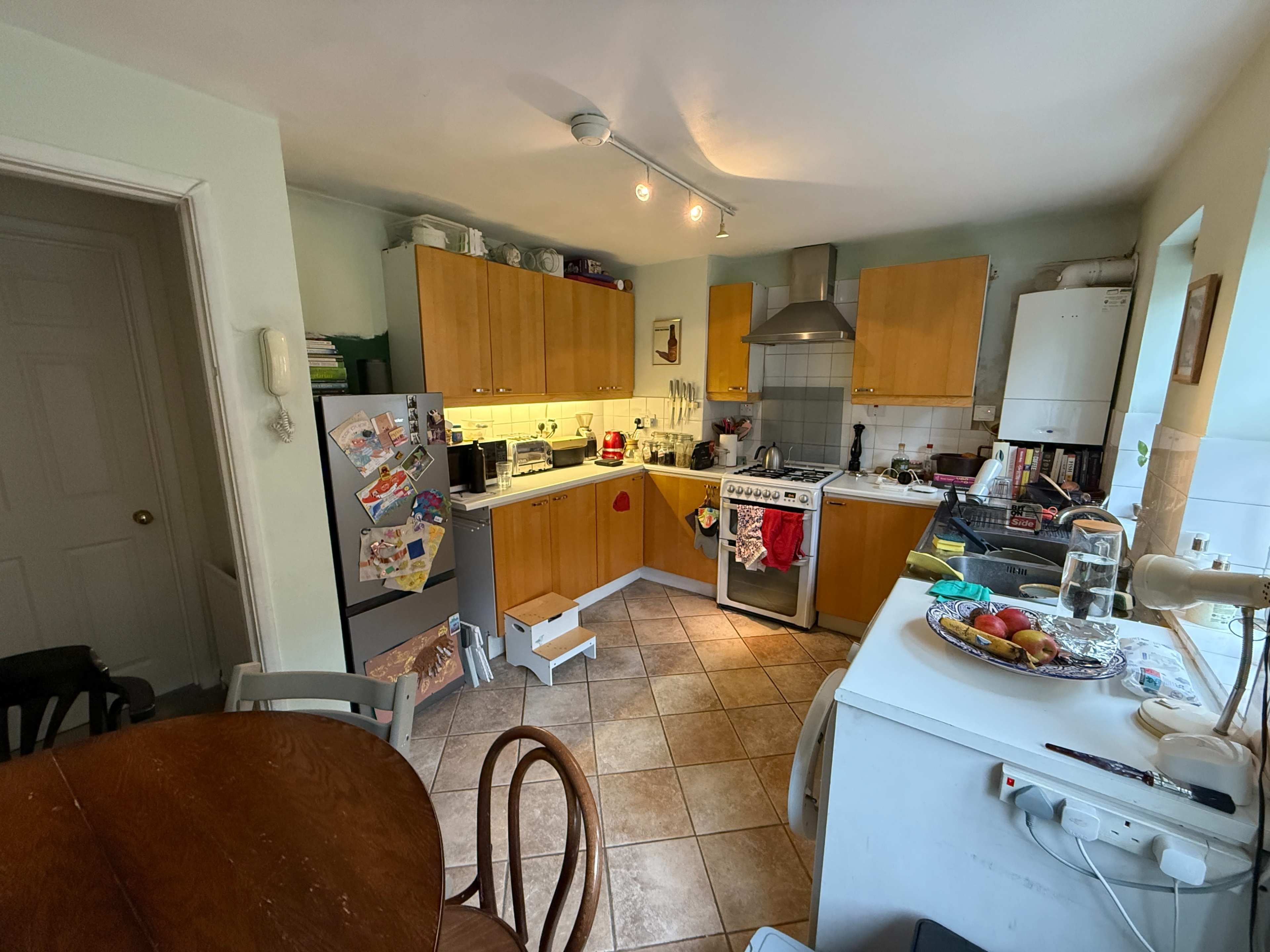 The image shows a kitchen featuring wooden cabinetry, a stove, a refrigerator covered in magnets and papers, and a dining table with chairs.