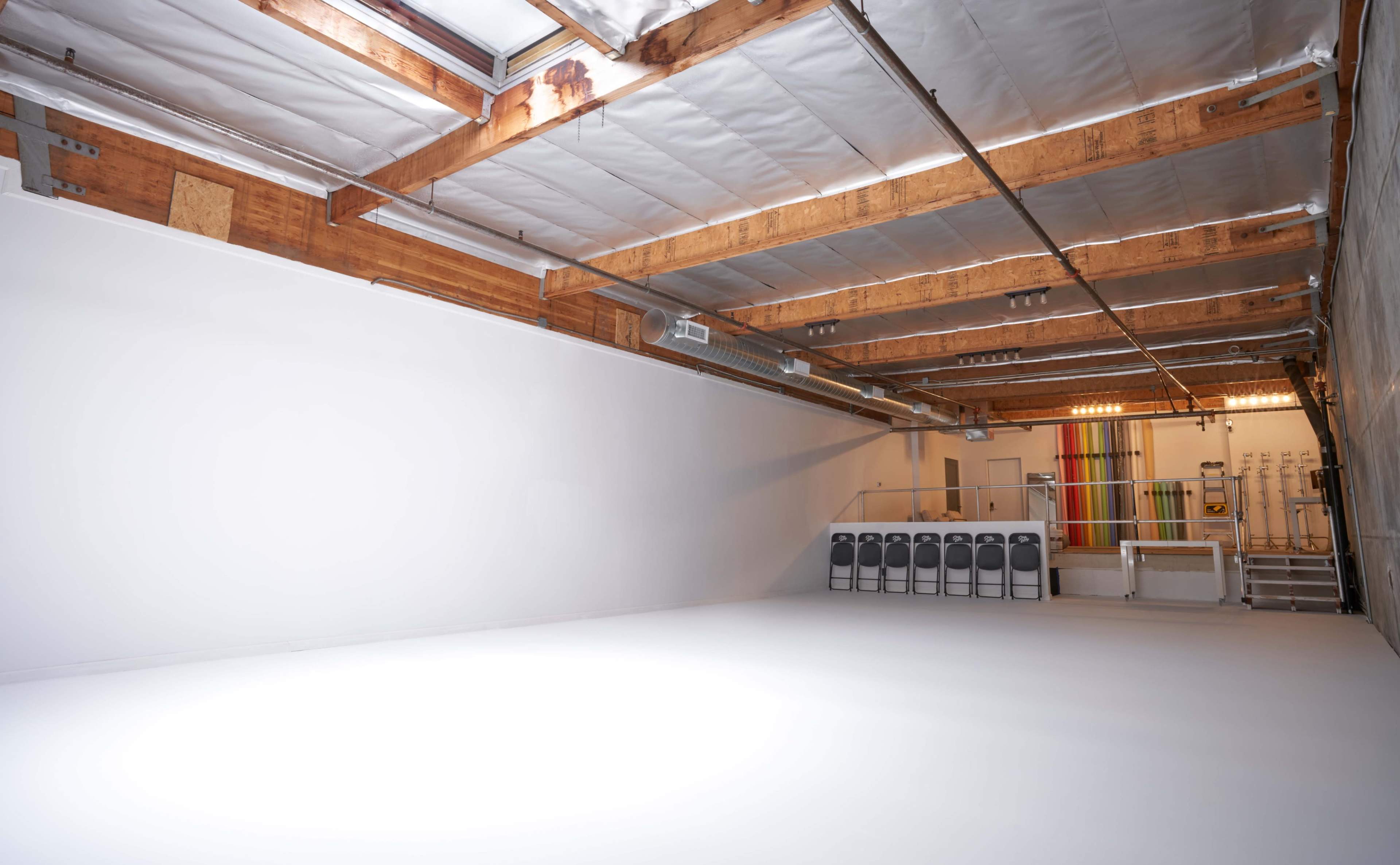 An empty studio space with a white floor, high wooden beams, and a wall designed for various backdrops.
