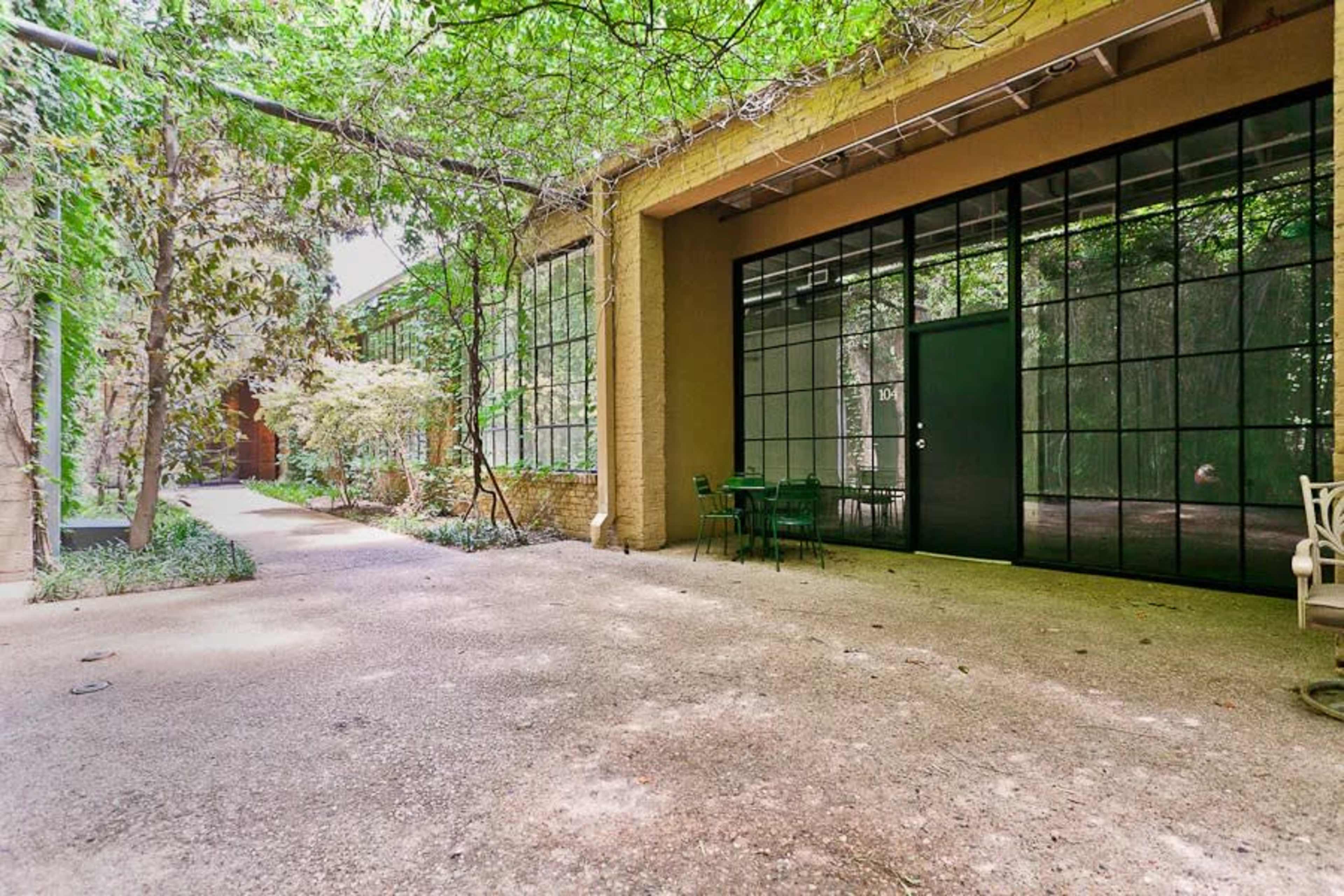 The image shows a serene outdoor space with a gravel pathway leading to a building featuring large glass windows and a dark door, surrounded by greenery.