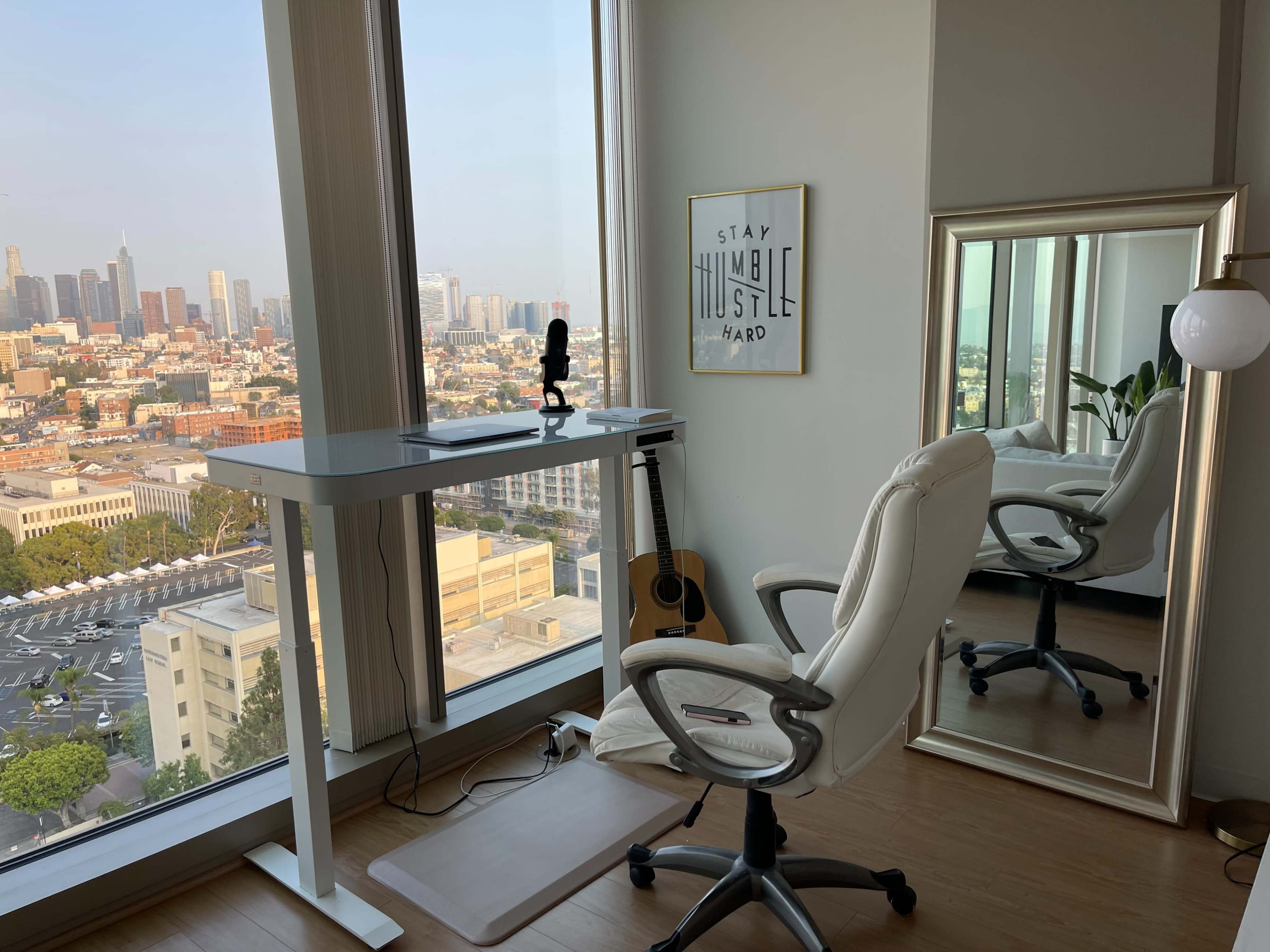 A modern workspace features a standing desk, a white chair, a microphone, a guitar, and a large window showcasing a city skyline.
